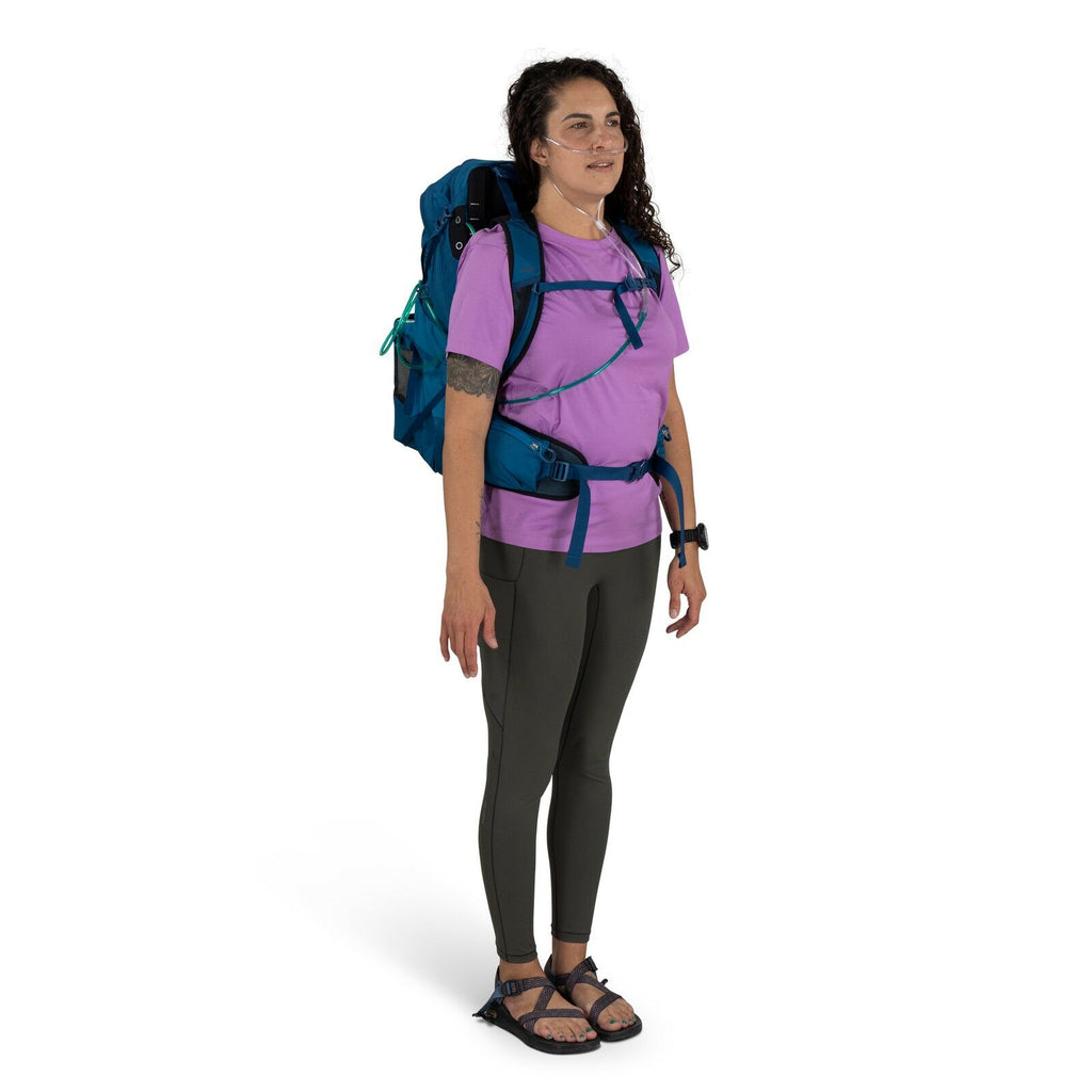 A person with curly hair wearing a purple shirt, brown leggings, and sandals stands with a blue backpack. They have nasal oxygen tubes, appearing relaxed.