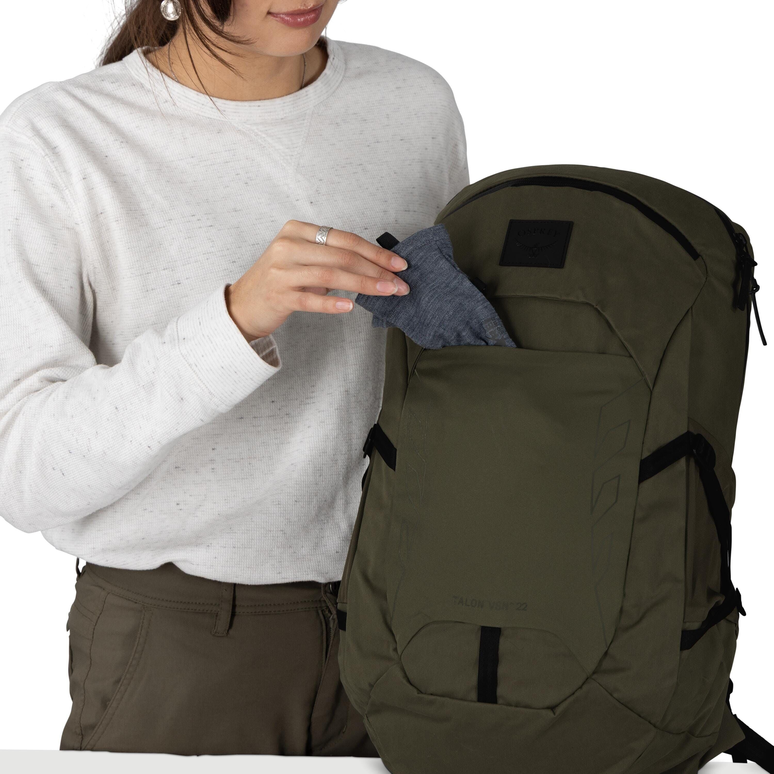A person wearing glasses and a light gray sweatshirt is holding an olive green Osprey backpack. They are slipping a pouch into the small top zip pocket. The backpack has a front panel with a black Osprey logo patch and side compression straps. The scene s