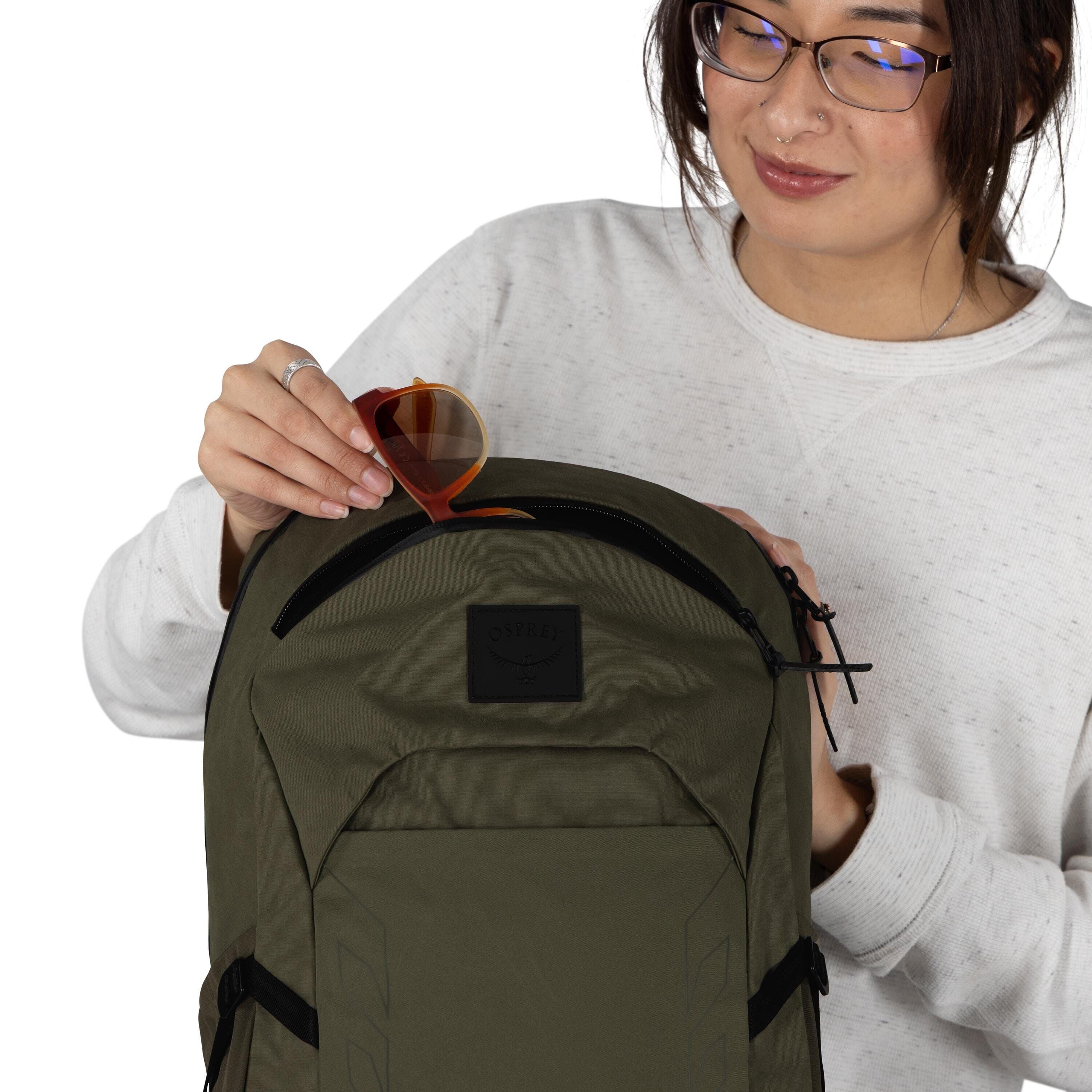 A person wearing glasses and a light gray sweatshirt is holding an olive green Osprey backpack. They are slipping a pair of orange-framed sunglasses into the small top zip pocket. The backpack has a front panel with a black Osprey logo patch and side comp