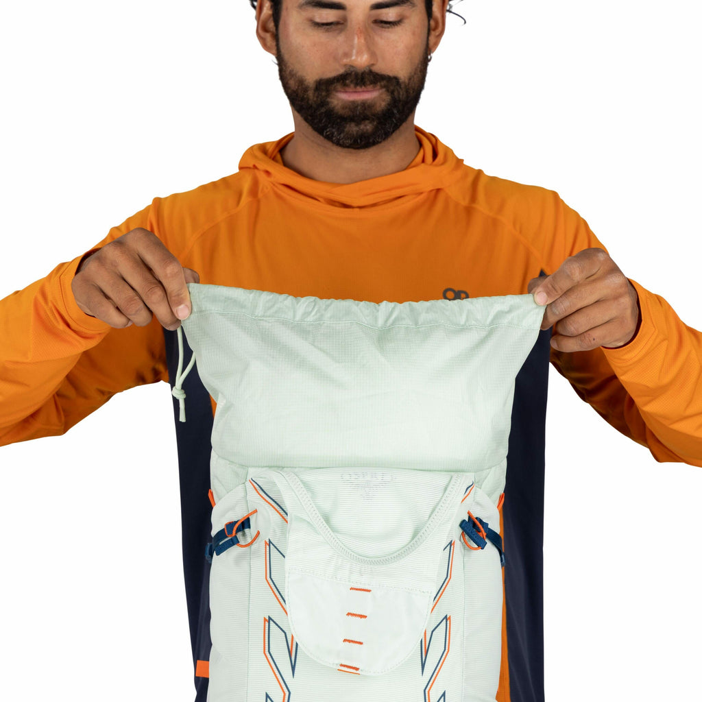 The image shows a person wearing an orange and navy long-sleeve shirt with the Outdoor Research (OR) logo on the left side of the chest. The individual is holding a light mint green backpack with a drawstring top, which appears to be made of a lightweight