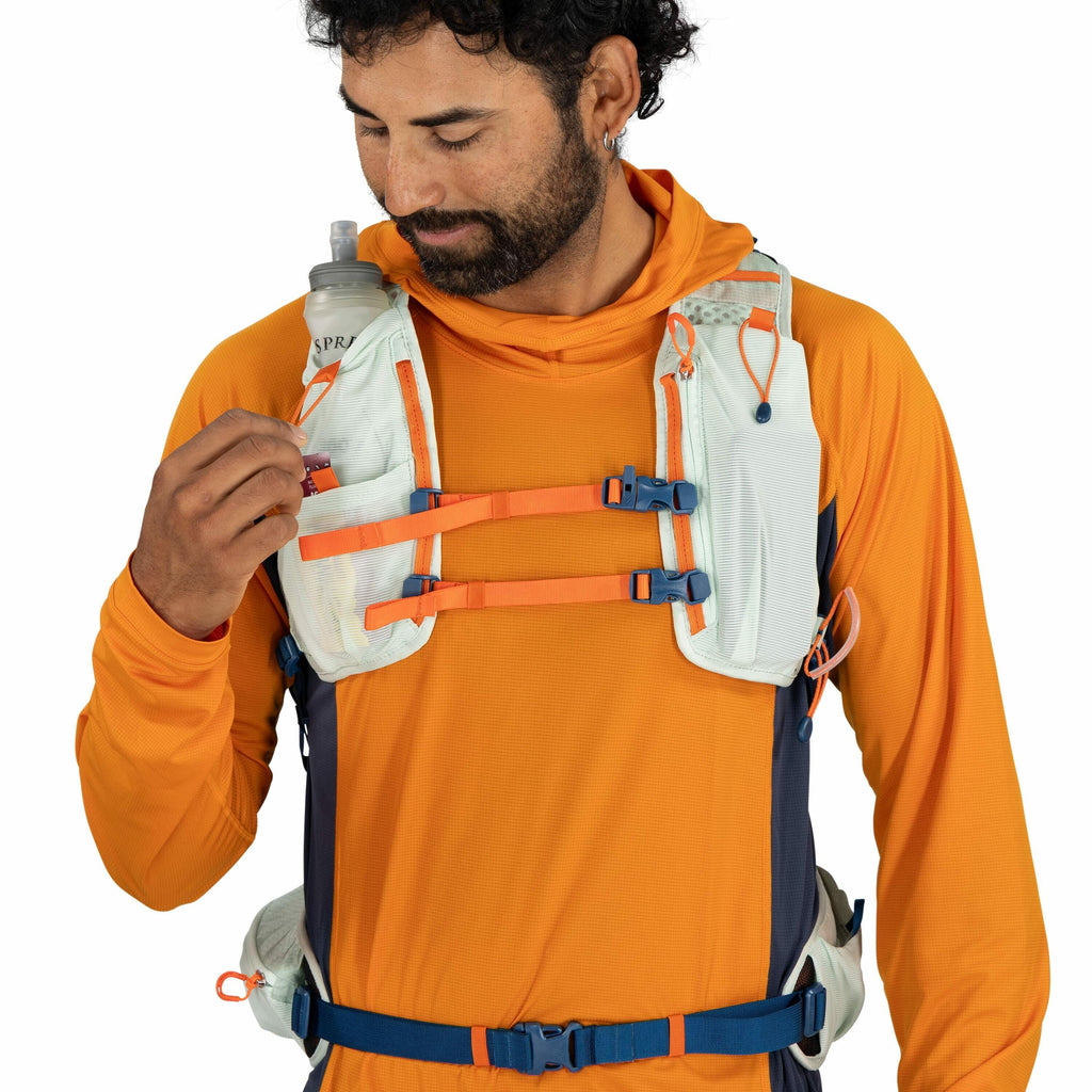 The image shows a person wearing an orange long-sleeve athletic shirt and a multi-colored hydration vest, which is primarily light blue and orange with dark blue accents. The vest features several adjustable straps across the chest, secured with blue and