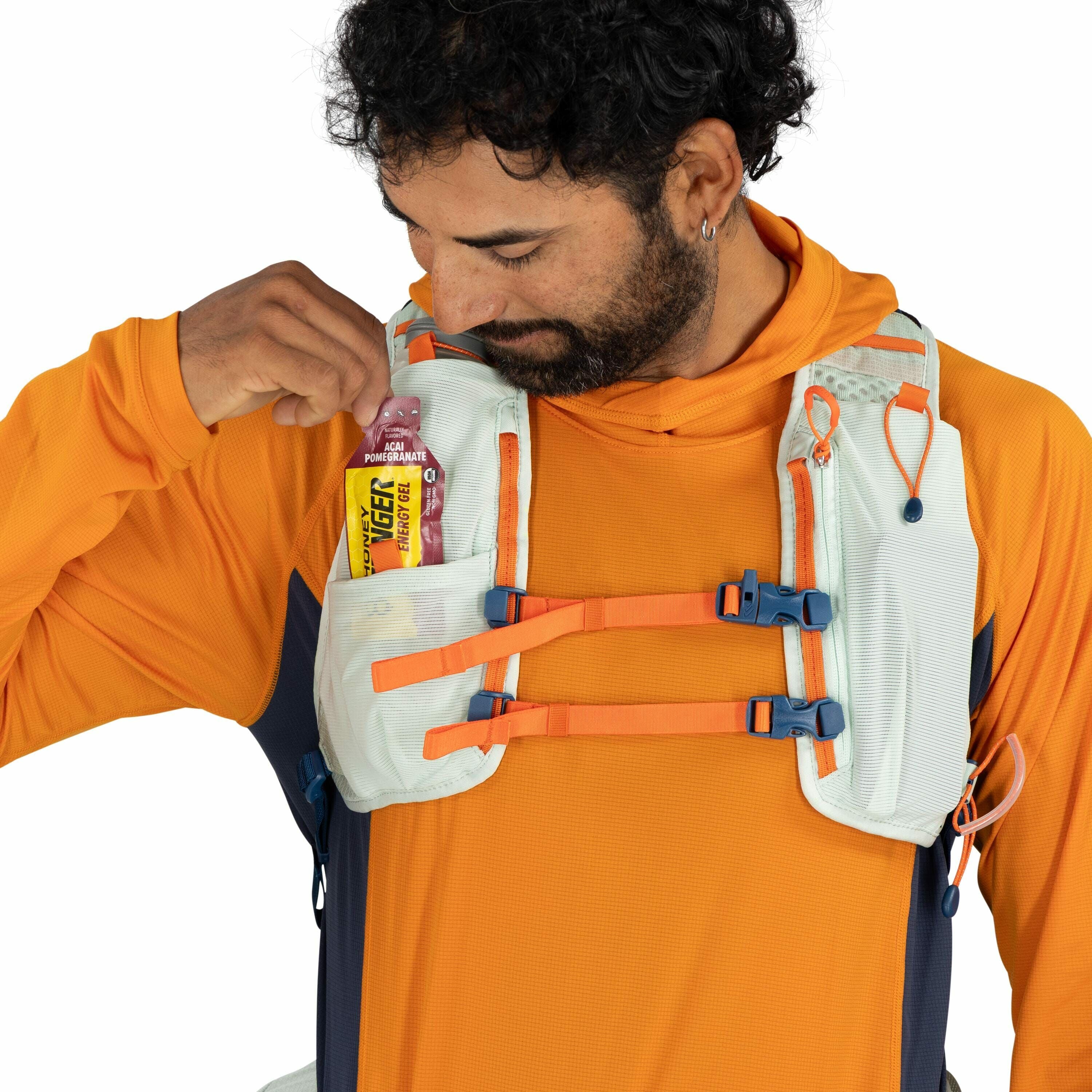 The image shows a person wearing an orange long-sleeve top and a white and orange hydration vest with blue accents. The individual is placing a Honey Stinger energy gel packet, labeled "Acai Pomegranate," into a pocket on the front of the vest. The vest h