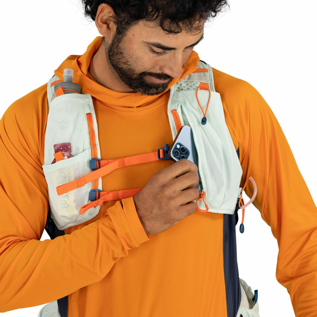 The image shows a person wearing an orange long-sleeve athletic top and a light-colored running vest with orange and blue accents. The vest has multiple pockets, with one containing a small water bottle and another holding a small snack. The person is see