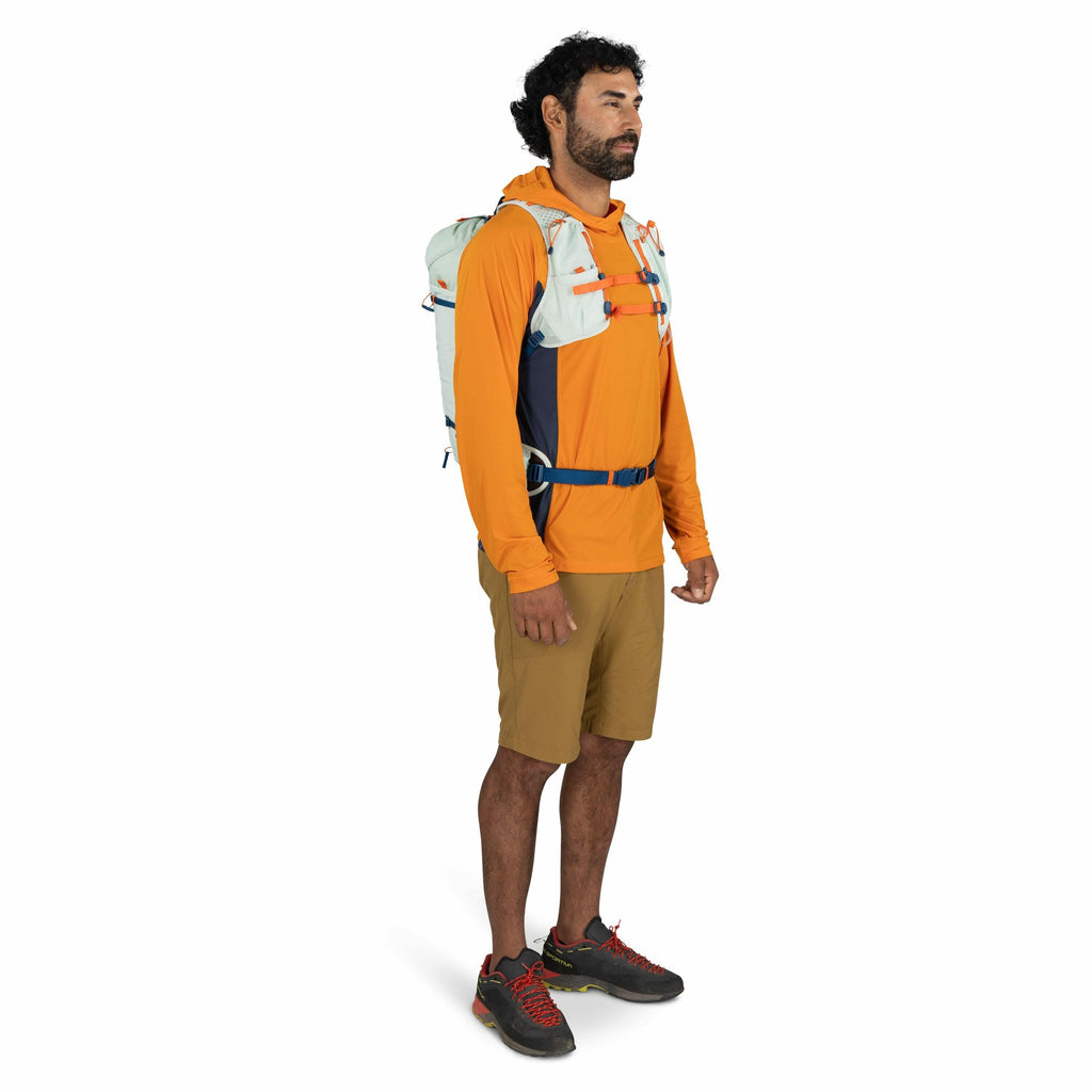 The image shows a person standing in a three-quarter profile view wearing outdoor and hiking attire. The individual is dressed in an orange long-sleeve shirt with a small white logo on the chest, a pair of light brown shorts, and black hiking shoes with r