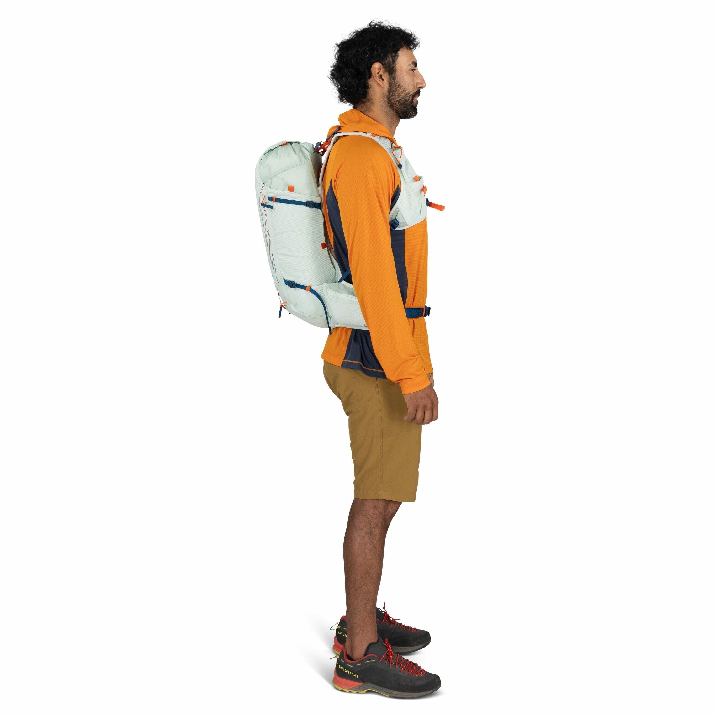 The image depicts a person standing in a side profile position, wearing outdoor clothing suitable for hiking or trekking. They are dressed in an orange and navy blue long-sleeve top, tan shorts, and grey hiking shoes with red laces. The individual is carr
