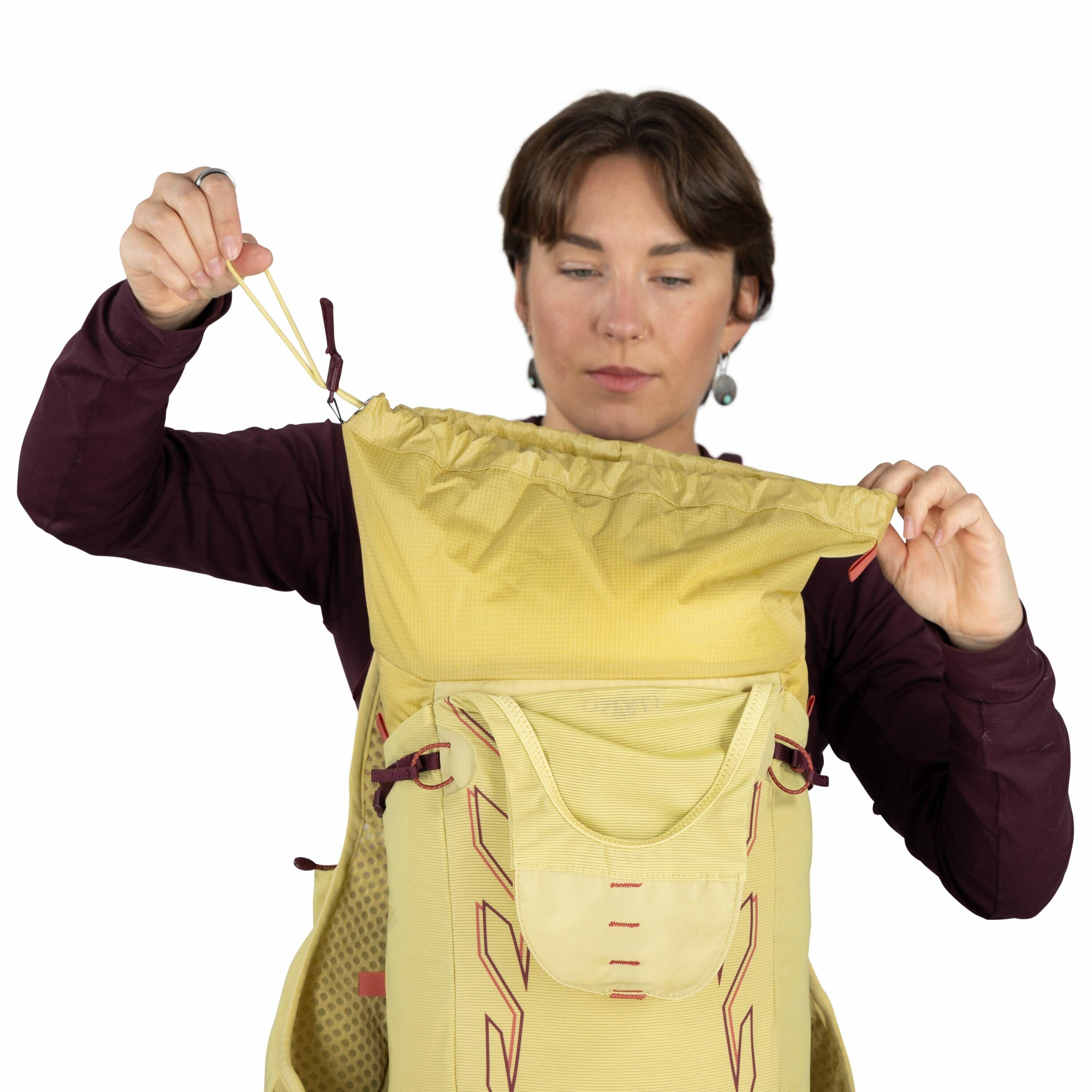 The image shows a person wearing a dark long-sleeve shirt, holding a large yellow backpack in front of them. The backpack has a roll-top closure, which the person is seen securing by pulling on the drawstring. It features a front pocket with red stitching