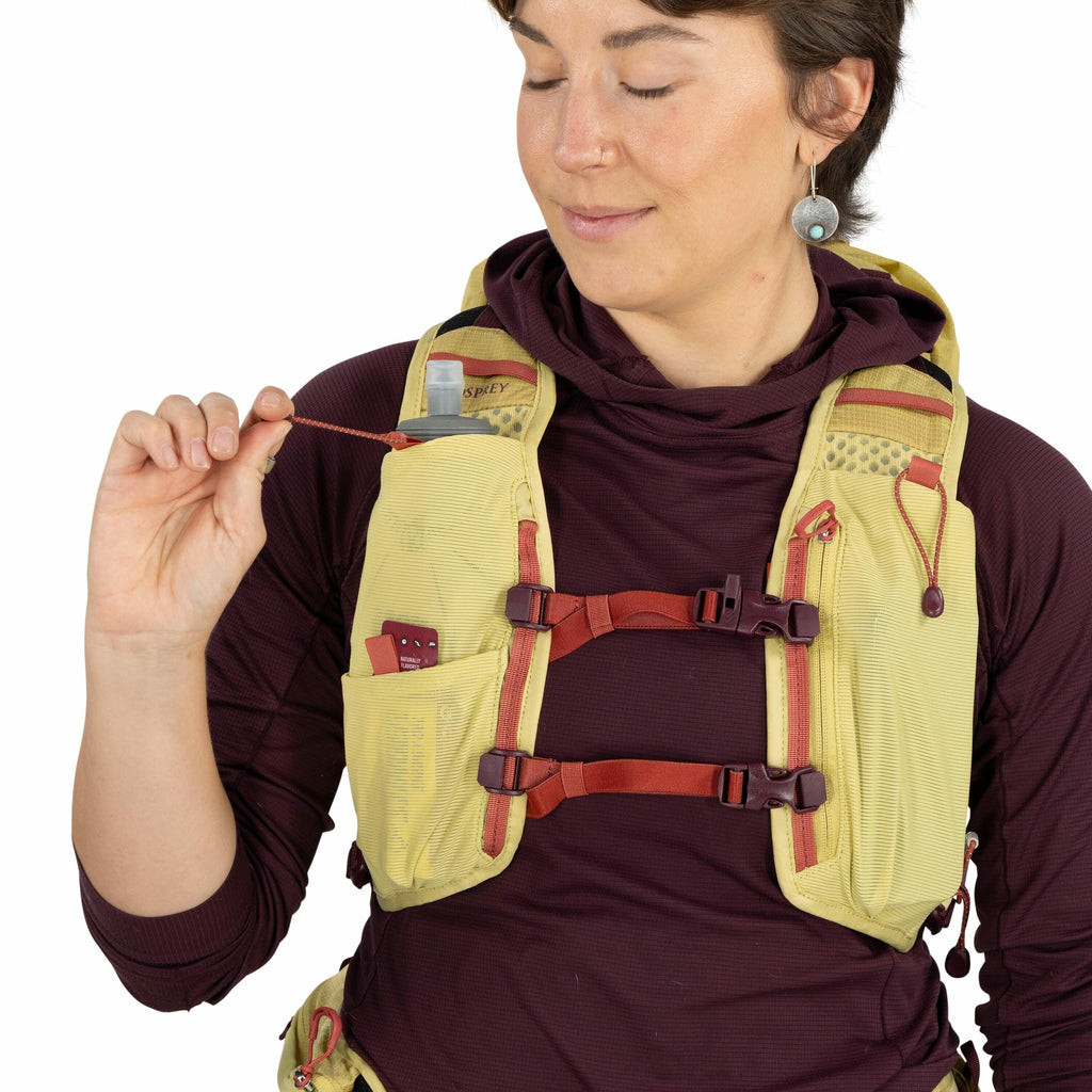 The image shows a person wearing an Osprey running vest in a light yellow color with red accents. The vest has multiple front pockets, one of which holds a small collapsible water bottle with a nozzle. Another pocket appears to contain a small item, possi
