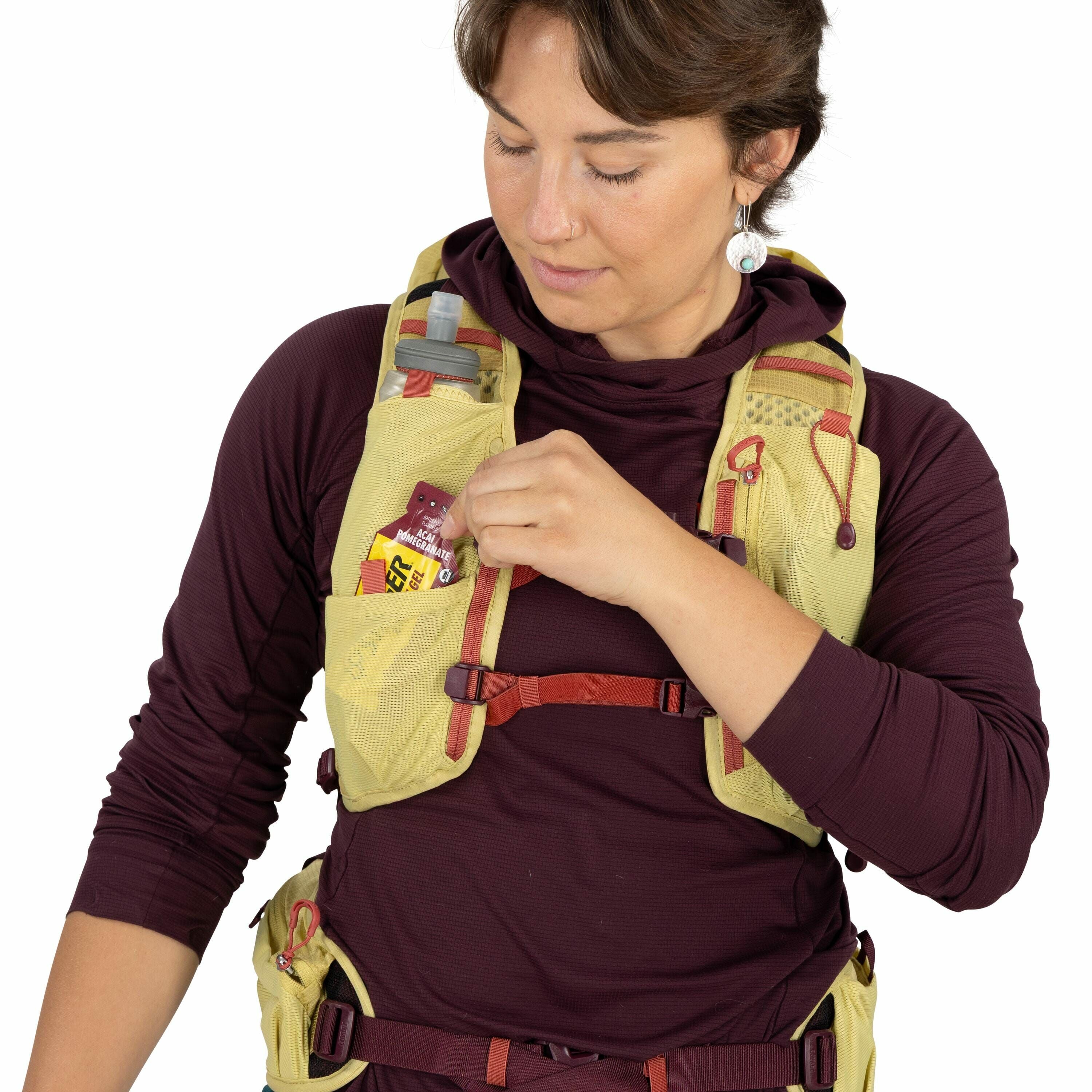 The image shows a person wearing a maroon long-sleeve athletic top and a yellow hiking or running hydration vest. The vest is equipped with various pockets and compartments, one of which contains a small water bottle with a red cap visible on the left sho