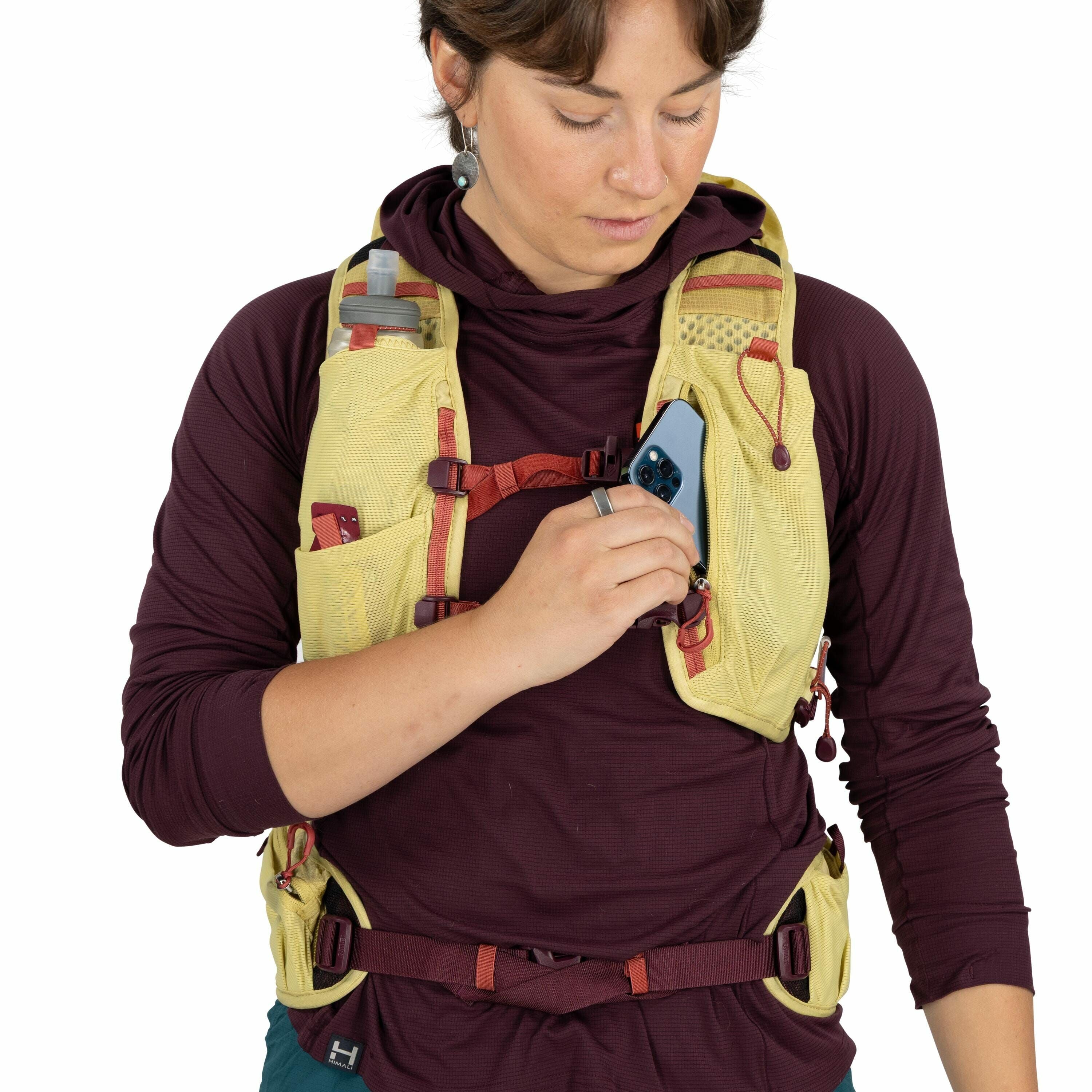The image shows a person wearing a yellow hydration backpack with adjustable straps and multiple compartments. One pocket is being used to store a smartphone, which the person is holding with their hand. Another pocket visibly contains a water bottle with