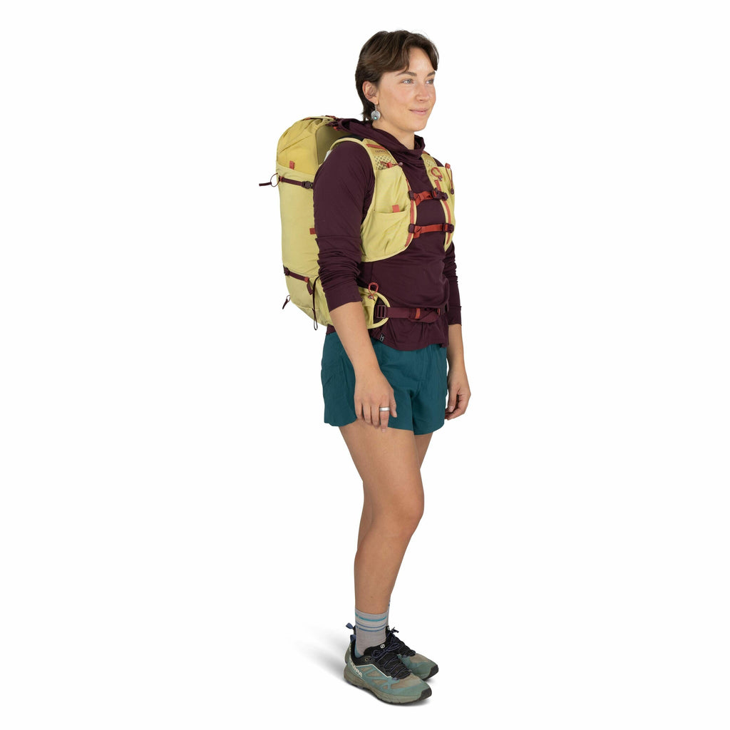 The image depicts a person standing and wearing outdoor hiking gear. The individual is equipped with a yellow backpack featuring red straps, which appear to provide a secure fit. They are dressed in a maroon long-sleeve shirt, teal shorts, and grey socks