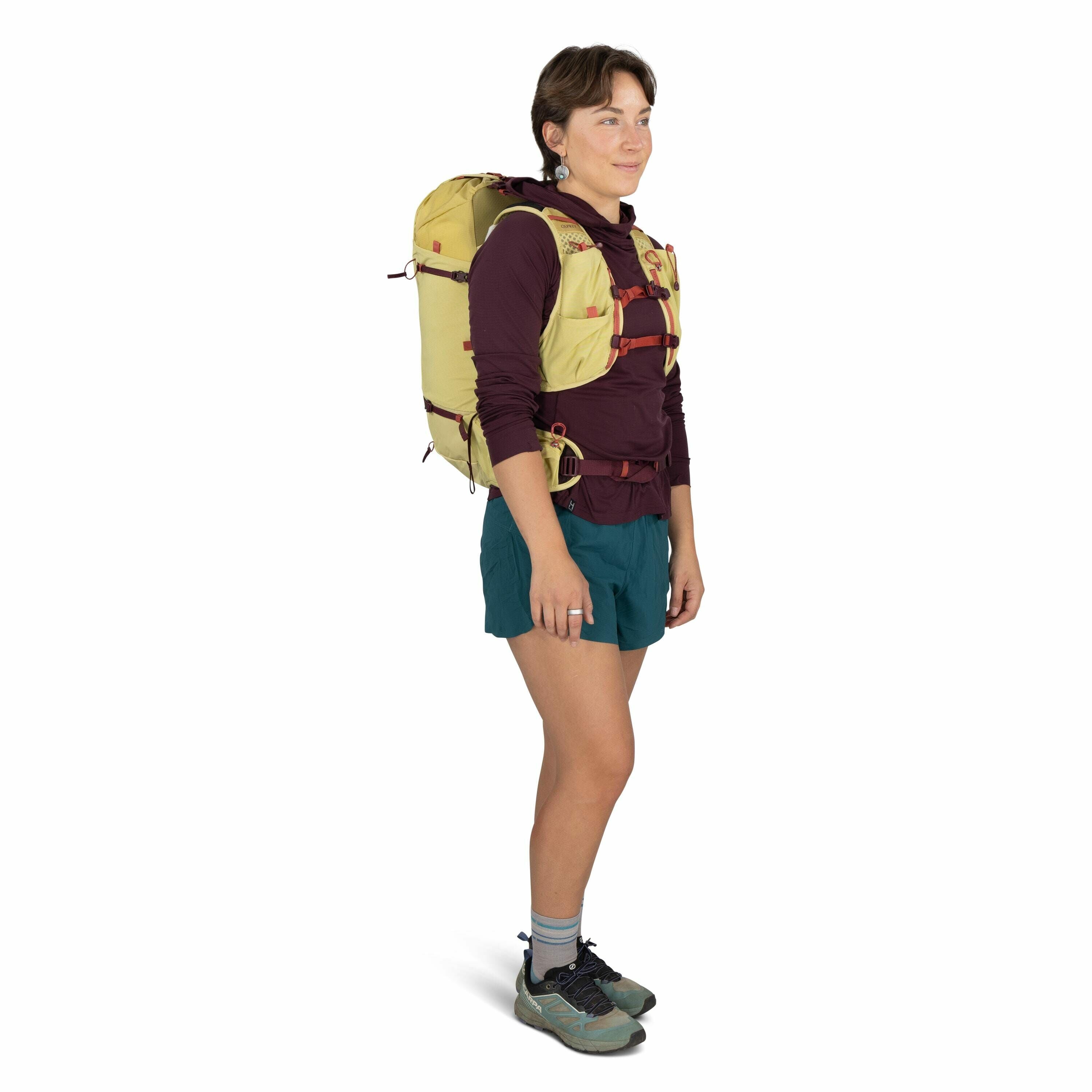The image depicts a person standing and wearing outdoor hiking gear. The individual is equipped with a yellow backpack featuring red straps, which appear to provide a secure fit. They are dressed in a maroon long-sleeve shirt, teal shorts, and grey socks