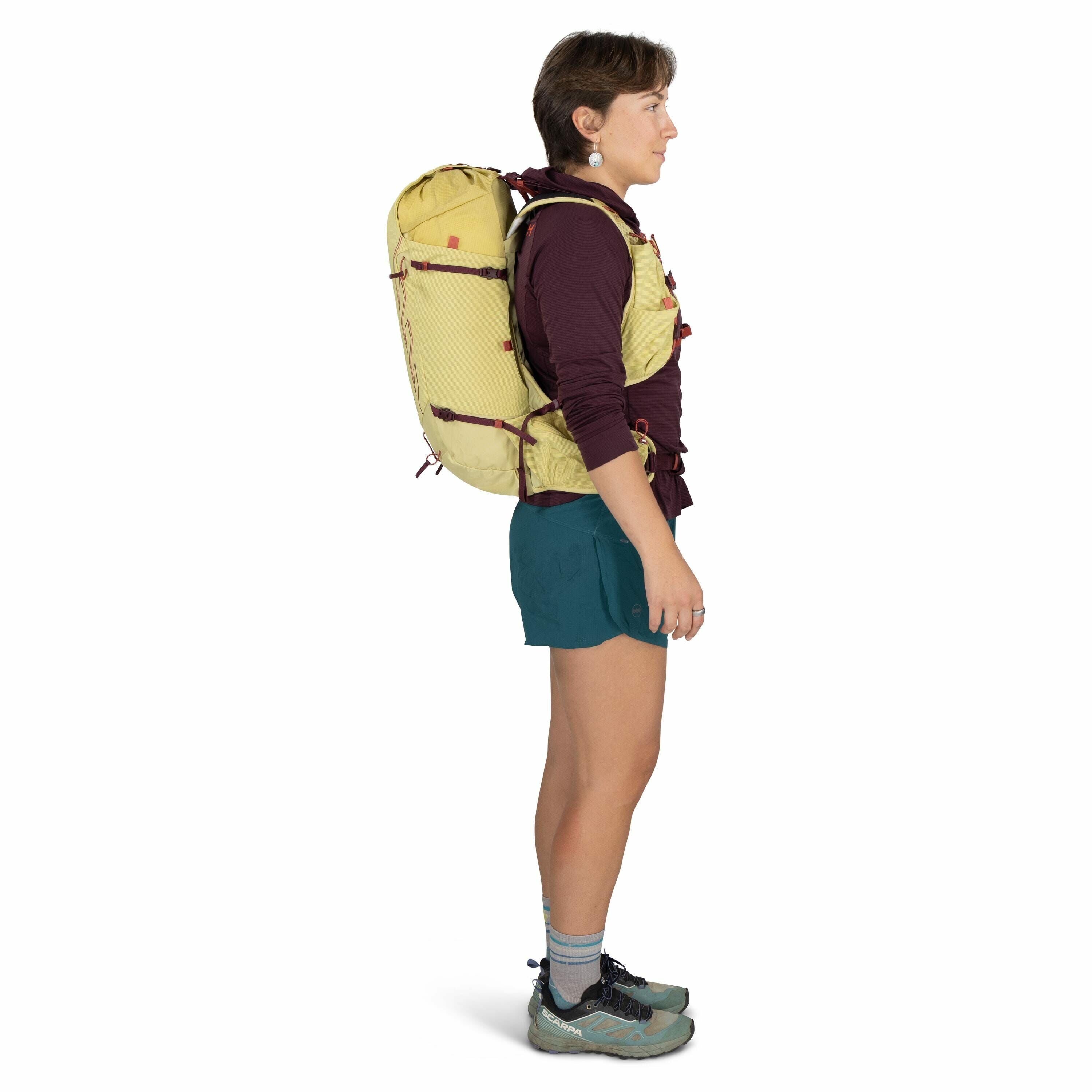 The image depicts a person standing sideways, dressed in hiking attire. They are wearing a burgundy long-sleeved top, teal shorts, and light blue socks with gray and teal trail running shoes from Altra. The person is carrying a large, yellow backpack with