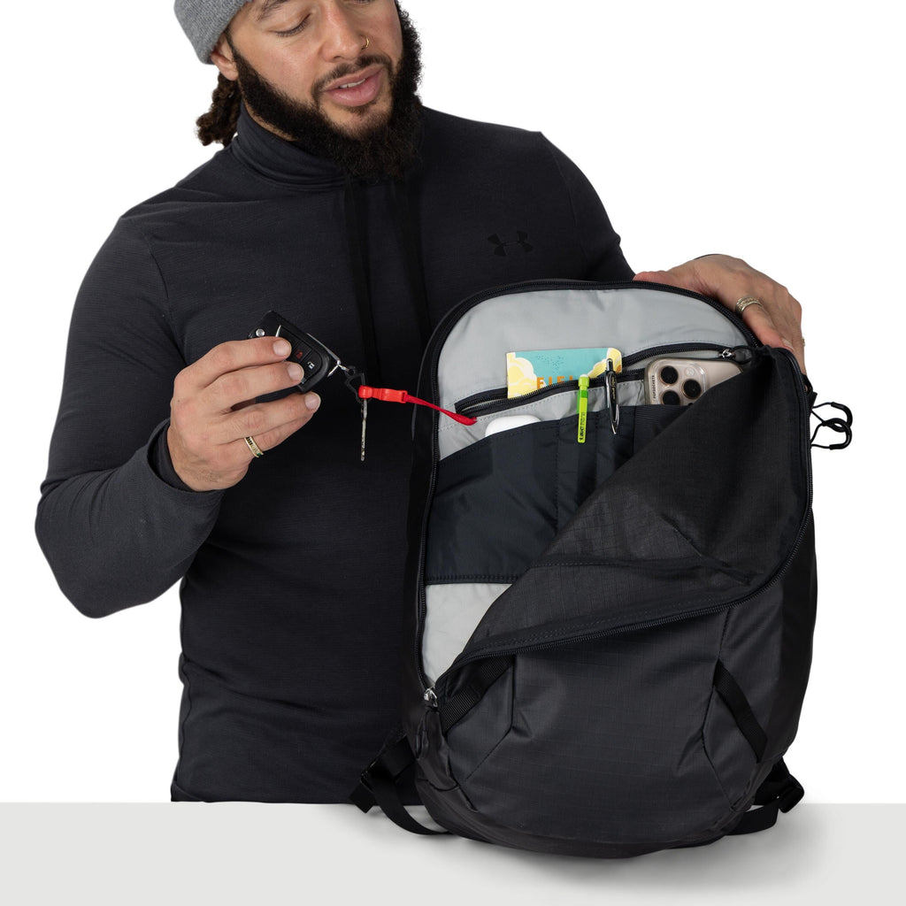 A person wearing a dark hoodie and a gray beanie is opening a black backpack and holding a tethered set of keys from the inside pocket. The pack's main compartment is unzipped wide, revealing an inner organizer pocket. The backpack has a sturdy, slightly