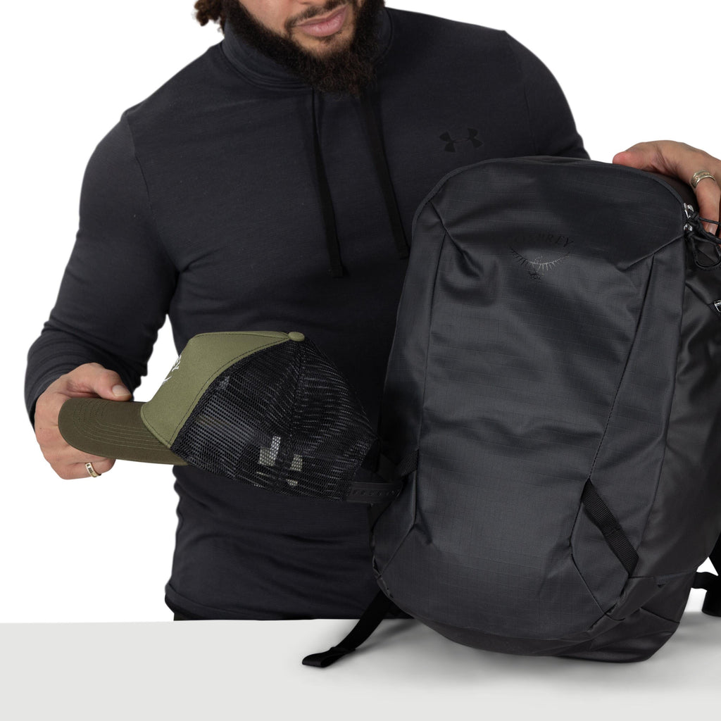 A person wearing a dark Under Armour pullover is holding a sleek black Osprey backpack. With the other hand, they're placing or removing an olive and black mesh trucker hat from the pack. The scene is shot against a plain white background with a table edg