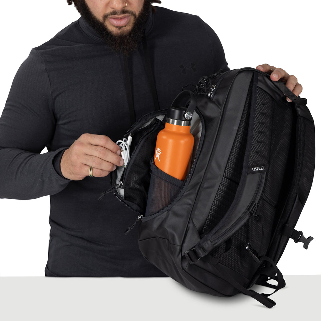 A person wearing a dark hoodie opens a black Osprey backpack. Inside, an orange Hydro Flask bottle sits in a mesh side pocket. The person is organizing a small zippered compartment, holding a white cable or earphones. The bag features padded straps and mu
