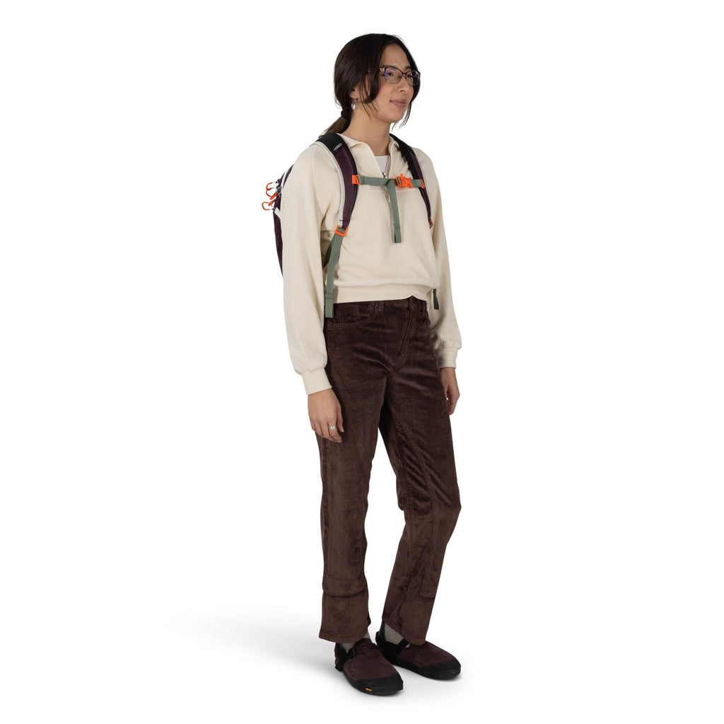A person stands in a casual outdoor outfit against a plain white background. They wear a cream sweatshirt, brown corduroy pants with a slight flare at the cuffs, and dark slip-on shoes. A backpack with gray straps and orange buckles is worn over the shoul