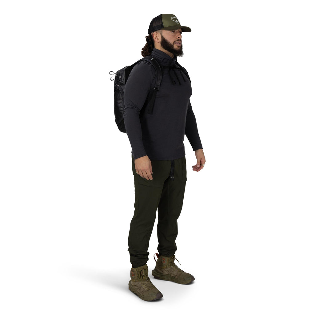 A person stands in a three-quarter pose wearing outdoor gear. They have on an olive baseball cap, a dark long-sleeve top, and olive jogger-style pants tucked into rugged olive hiking boots. A black backpack is strapped on their shoulders, and a neck gaite