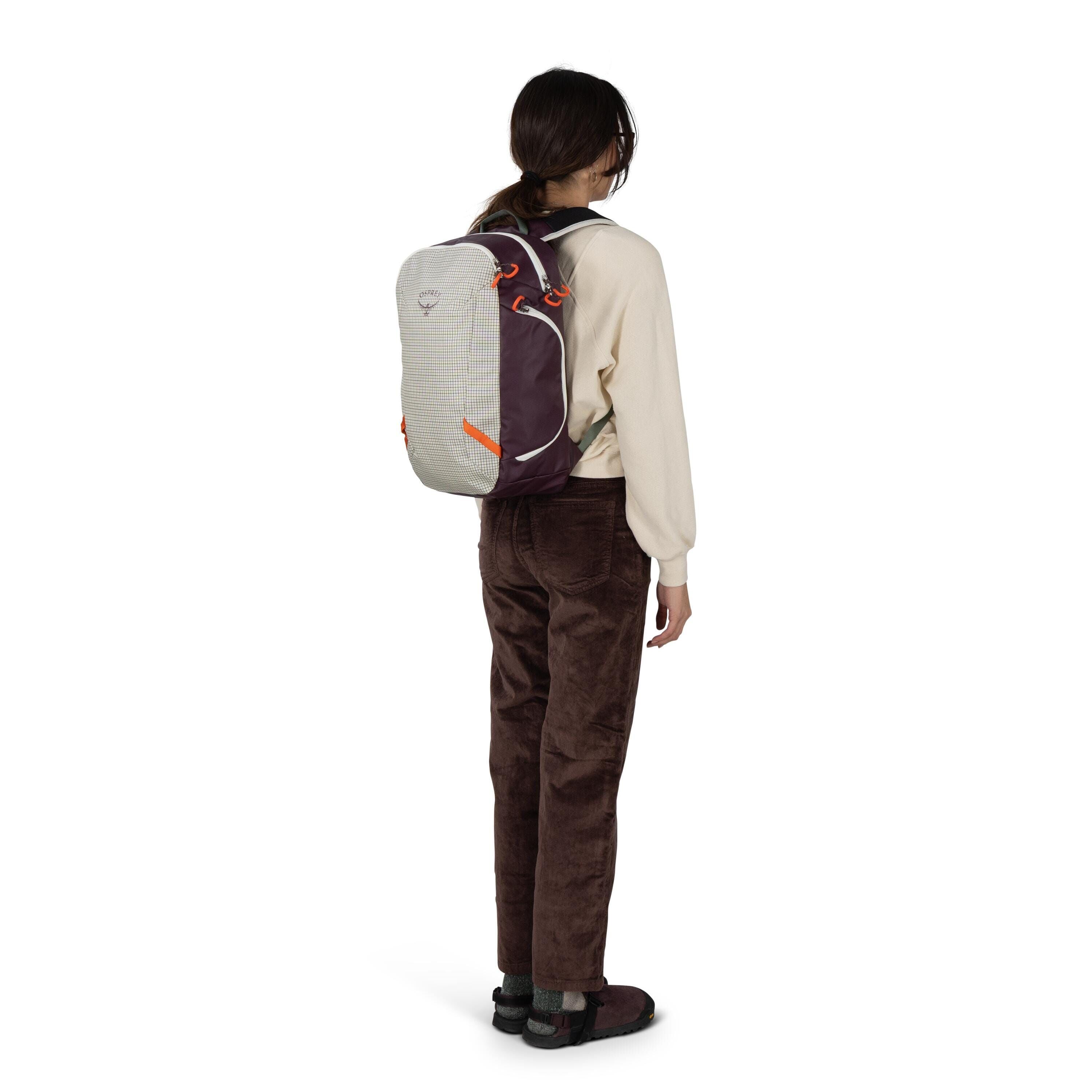 A person stands facing away, wearing a compact Osprey backpack with a light gray, grid-textured front panel and dark maroon sides accented by orange loops and zipper pulls. They have on a cream sweatshirt, brown corduroy pants, and dark slip-on shoes with