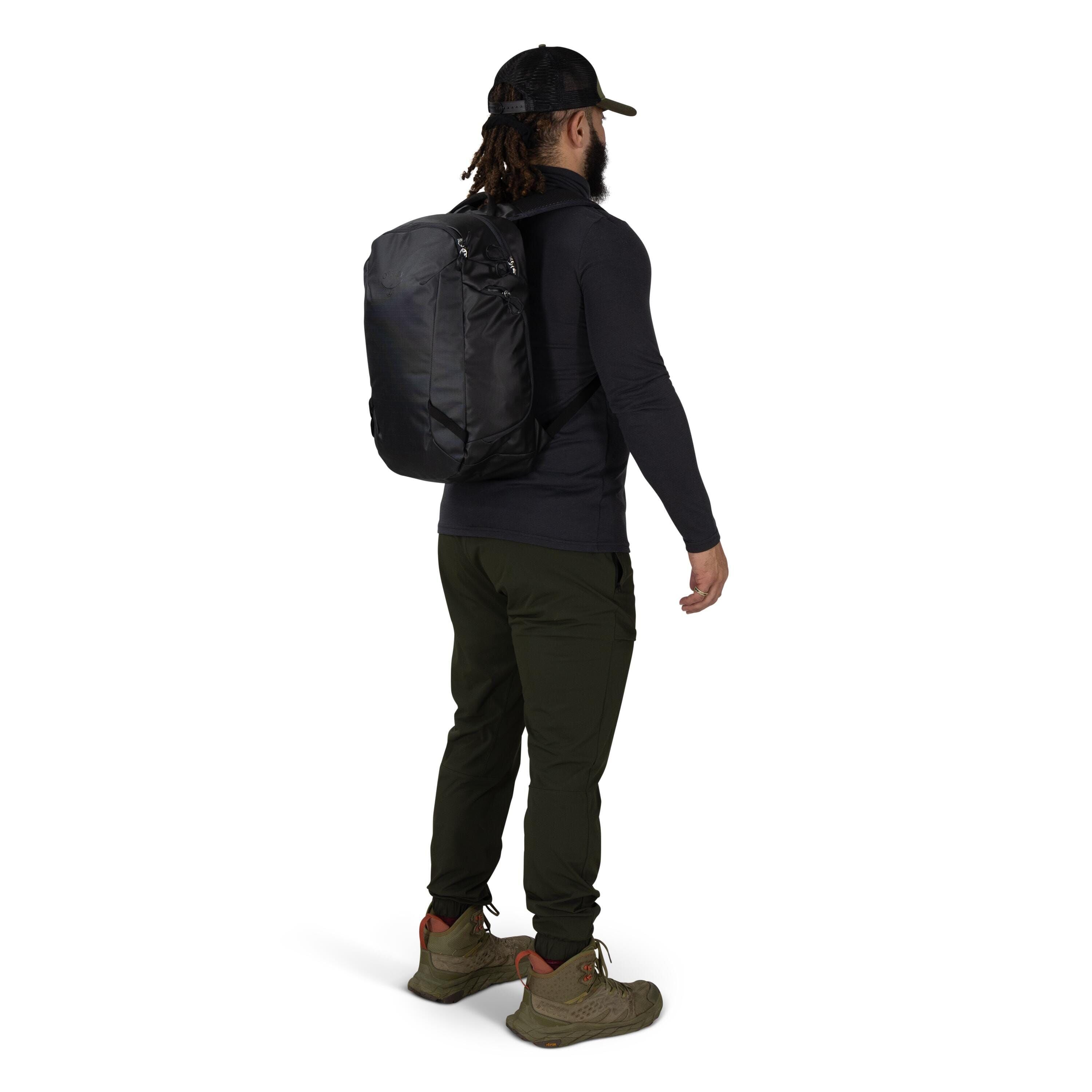 A person is shown from the back wearing a black backpack. They're dressed in outdoor attire: a dark long_sleeve top, olive green pants, and olive hiking boots with orange accents. The person also wears a black mesh-back cap. The background is plain white,