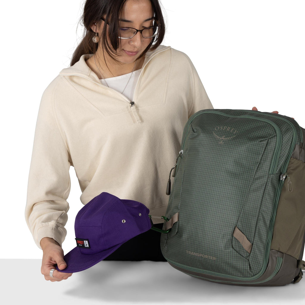 A person wearing glasses and a cream quarter-zip sweater is holding a green Osprey backpack and a purple cap. The cap is clipped to the backpack with a small carabiner. The backpack has a textured front panel with the Osprey logo and the word ?Transporter