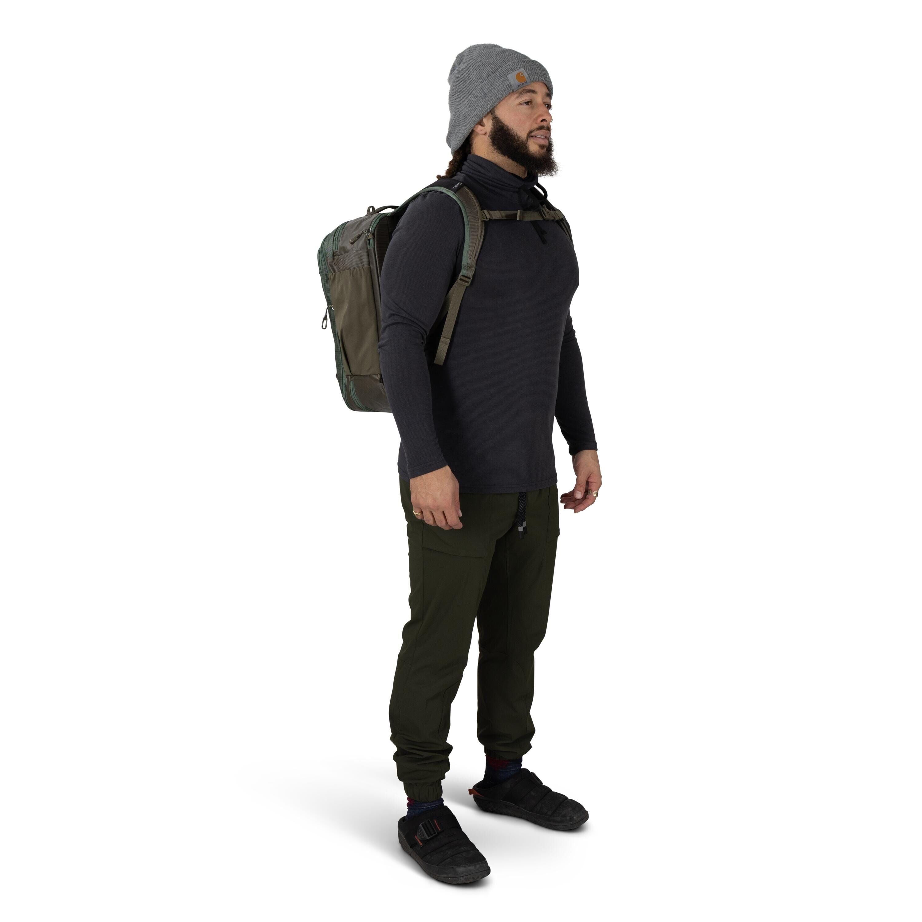 A person is standing in a side-facing pose wearing outdoor clothing and a gray beanie. They have on a dark long-sleeve top, olive-green jogger-style pants, and black shoes with strap closures. An olive backpack with padded shoulder straps and a sternum st