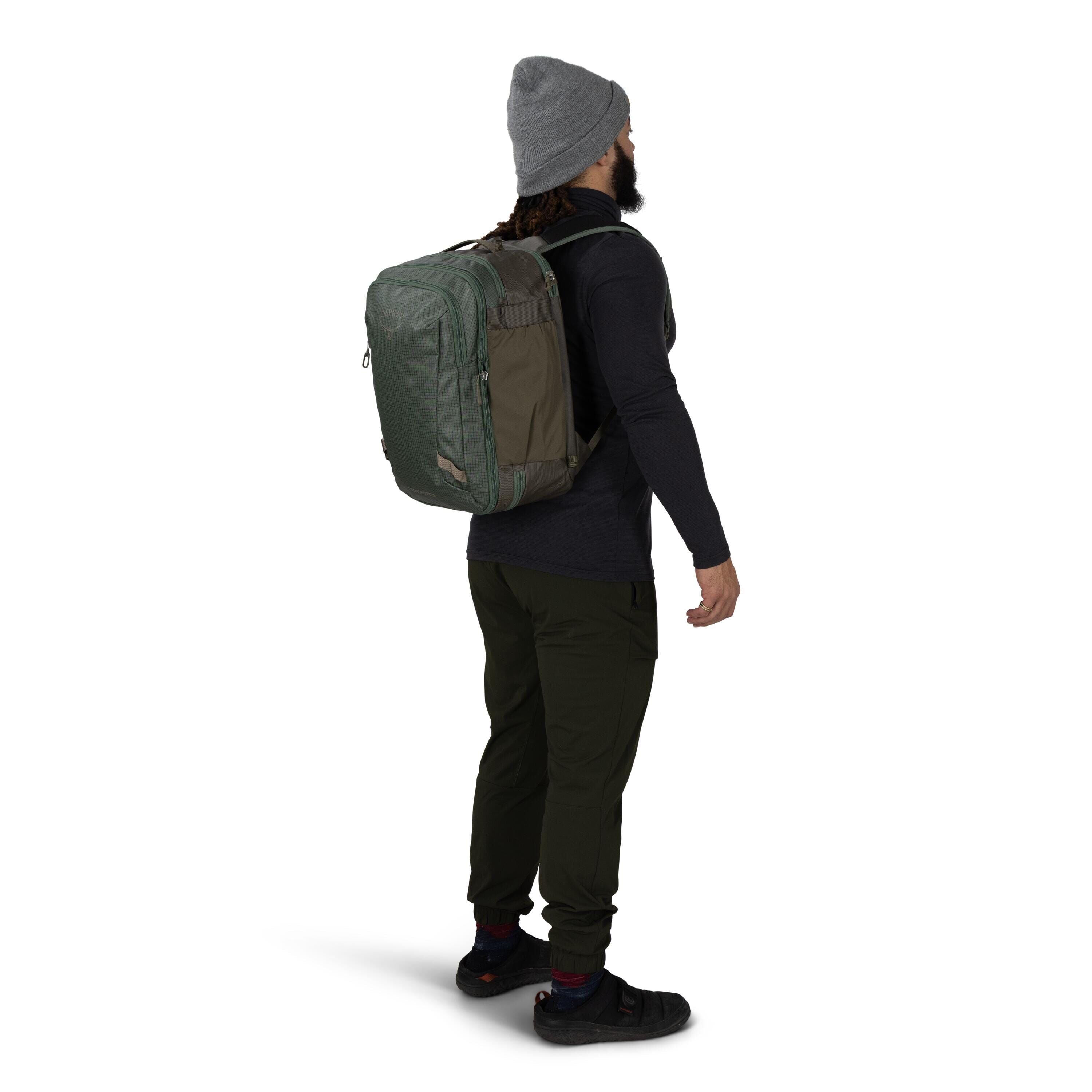 A person wearing outdoor clothing stands in profile, carrying a large green and olive backpack. They have on a gray beanie, a dark long_sleeve top, dark green pants, and black shoes with visible red socks. The backpack appears structured with multiple zip