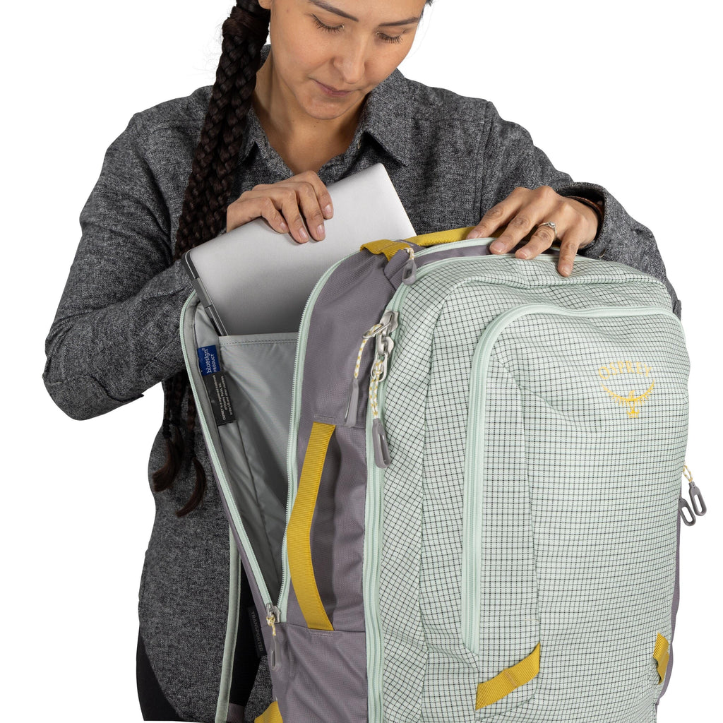 The image shows a person inserting or removing a laptop from the main compartment of a backpack. The backpack is predominantly light green with a checkered pattern and has light grey accents and yellow straps. It is designed with a large front zippered co