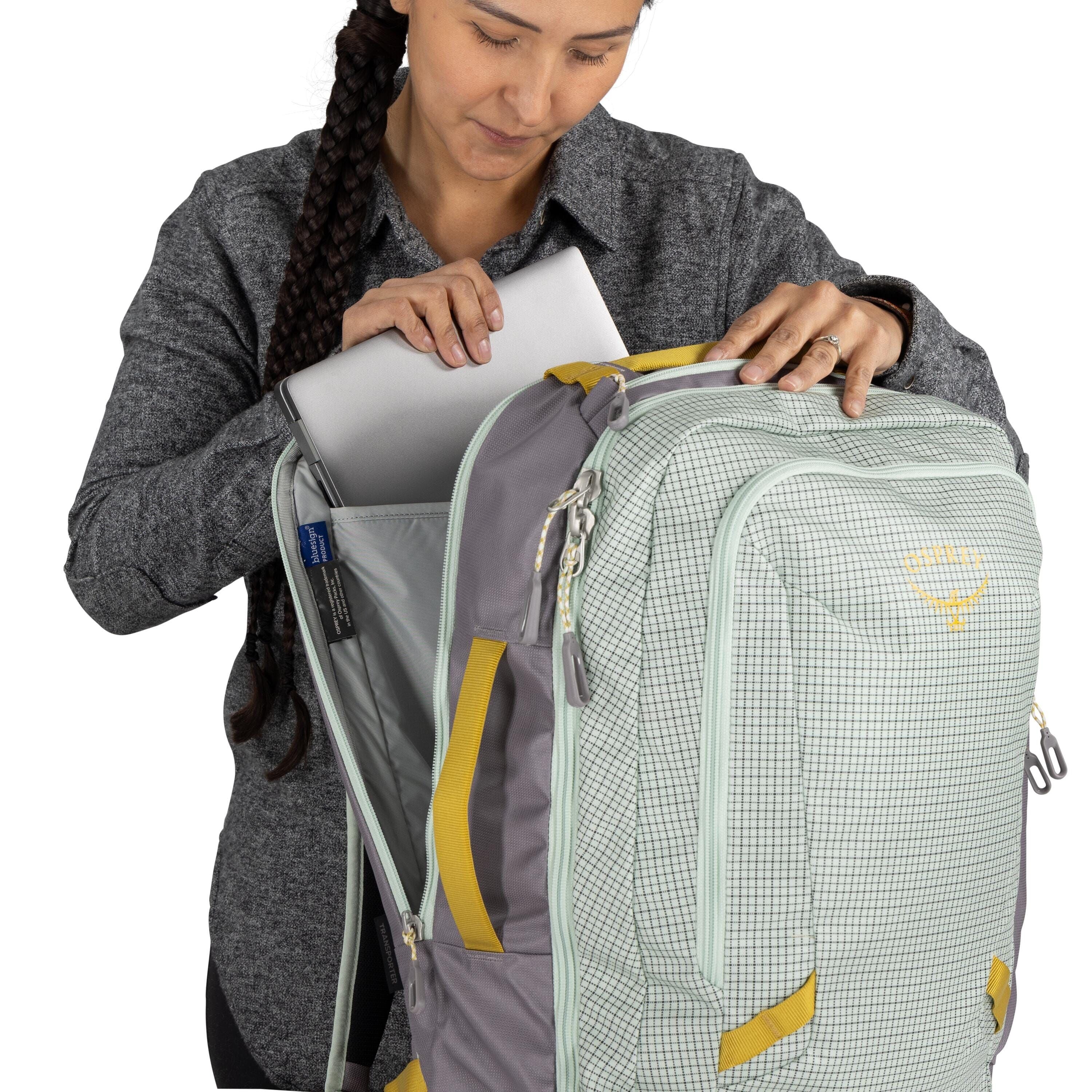 The image shows a person inserting or removing a laptop from the main compartment of a backpack. The backpack is predominantly light green with a checkered pattern and has light grey accents and yellow straps. It is designed with a large front zippered co
