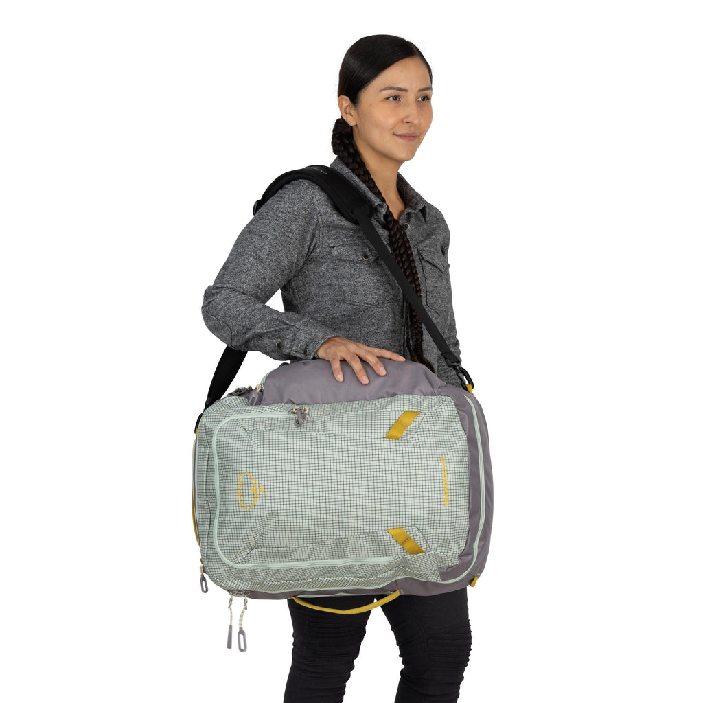 The image depicts a person standing and holding a bag with a shoulder strap, worn across the body. The individual is wearing a gray button-up shirt and black pants. The bag is primarily light gray with a checked pattern and features yellow accents, such a