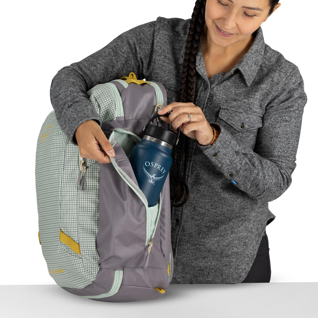 The image shows a person holding a backpack and placing a blue Osprey water bottle into one of its zippered pockets. The backpack is a combination of mint green with a checkered pattern and gray with yellow accents. The person is dressed in a gray button-