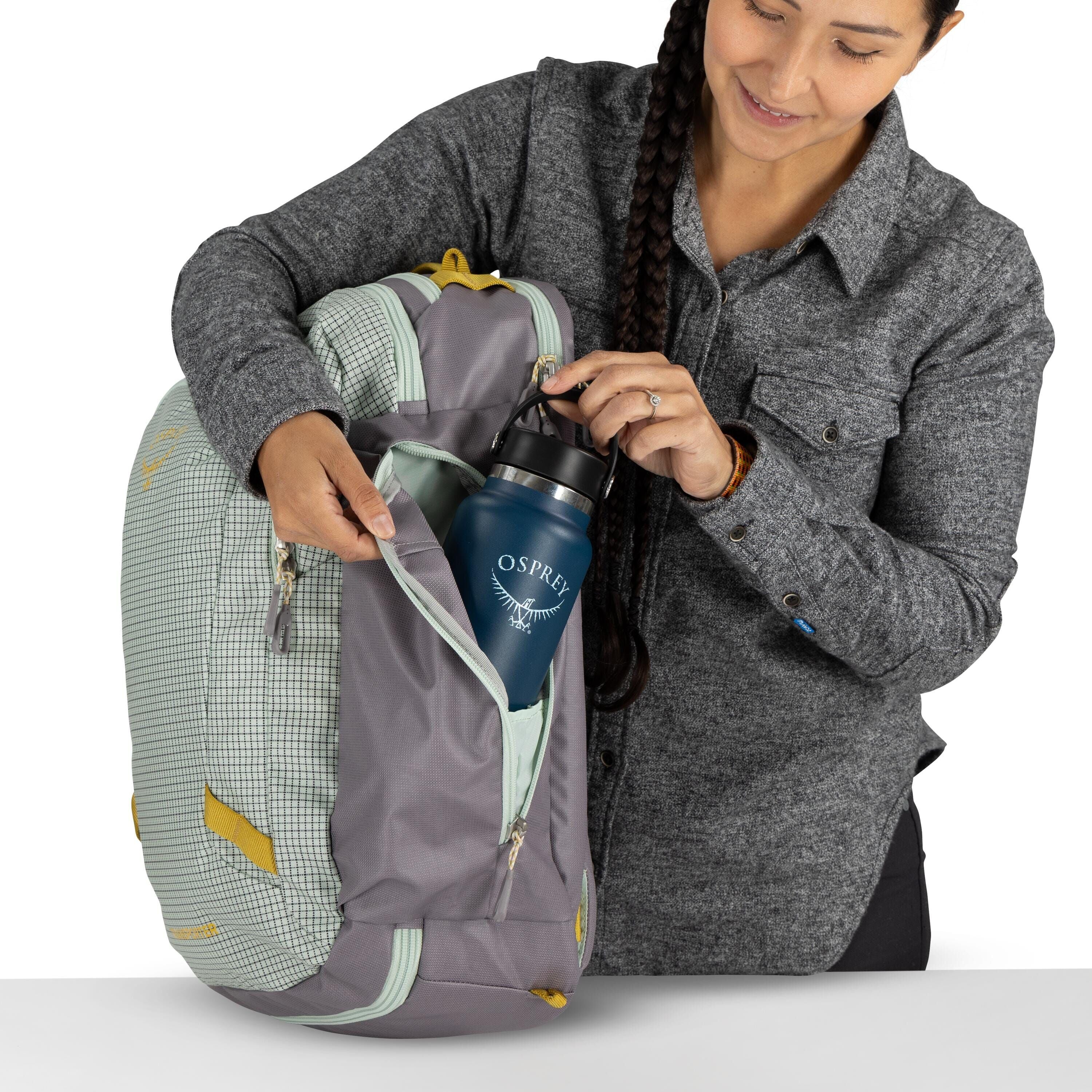 The image shows a person holding a backpack and placing a blue Osprey water bottle into one of its zippered pockets. The backpack is a combination of mint green with a checkered pattern and gray with yellow accents. The person is dressed in a gray button-