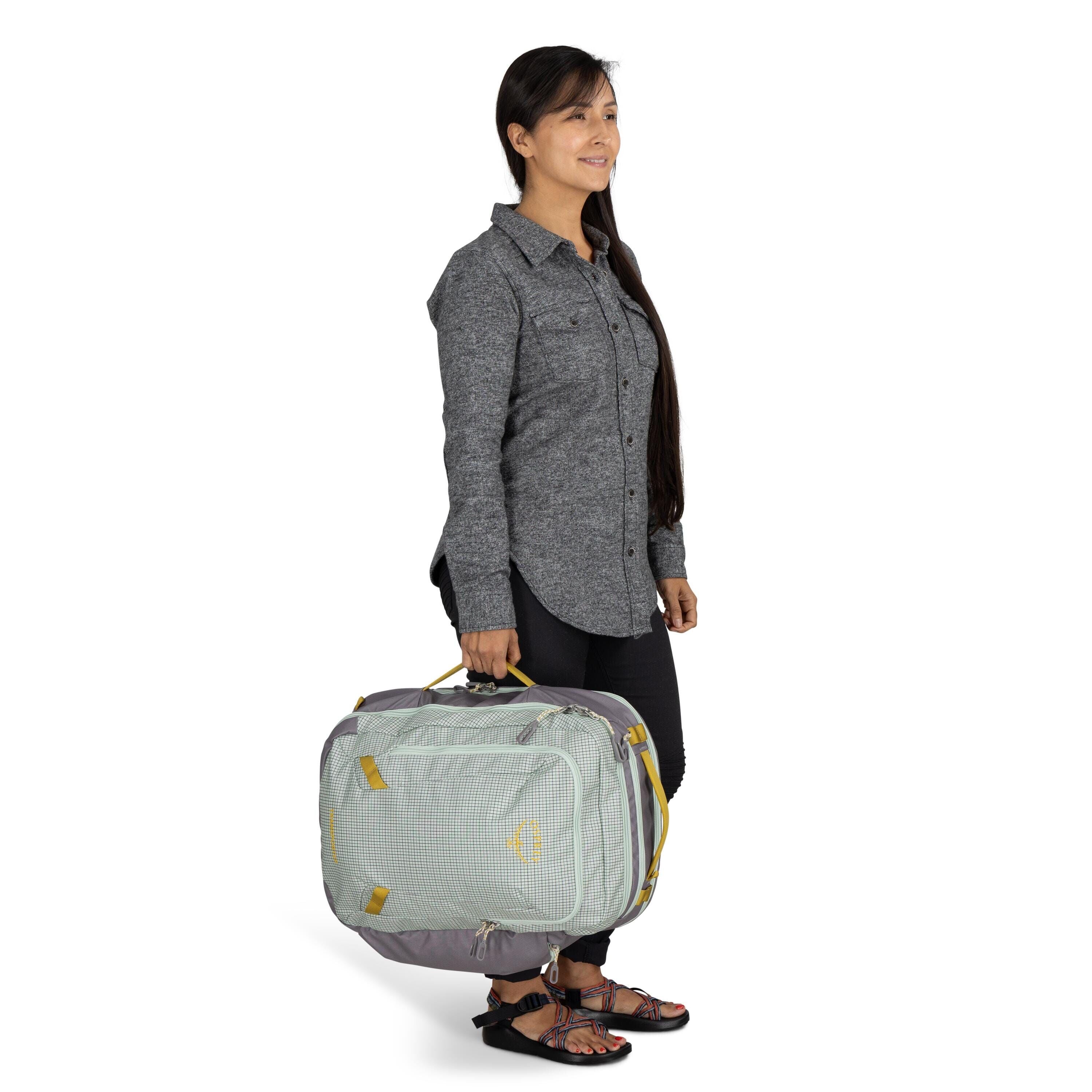 The image features a person standing and holding a soft travel bag. The individual is dressed in a long-sleeved, buttoned-up gray shirt and black pants, paired with black sandals. The bag they are holding is a light shade of green, with yellow accents and