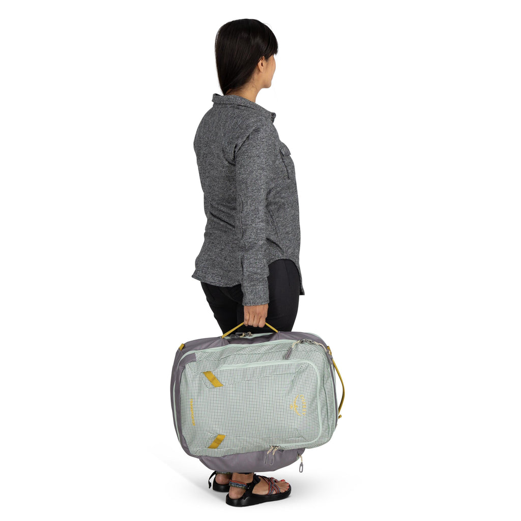 The image shows a woman standing and holding a large, light-colored travel backpack by its side handle with one hand. The backpack features a grey and green color scheme with yellow accents. She is wearing a grey long-sleeved sweater, black pants, and bla