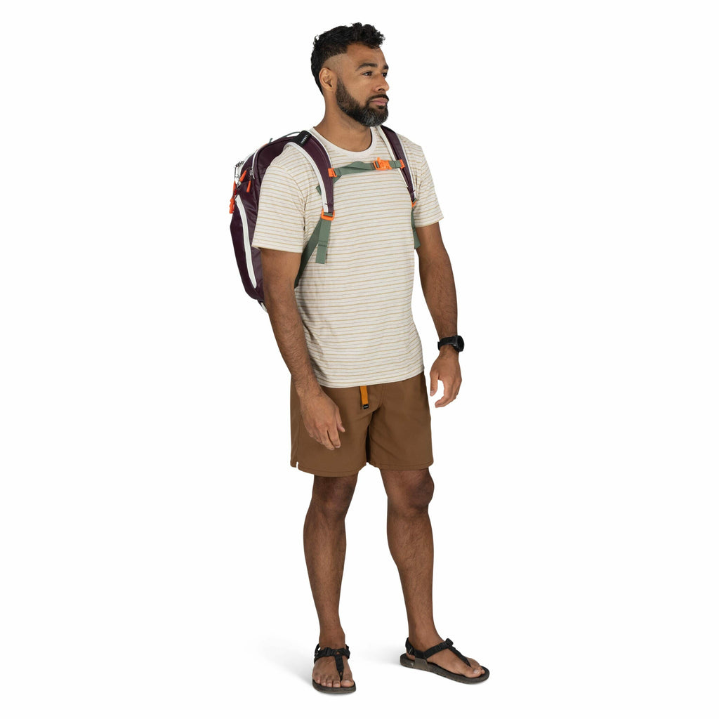 The image features a person standing against a plain white background, dressed in casual outdoor attire. The individual is wearing a light-colored, short-sleeved, striped t-shirt paired with brown shorts. They have on black sandals and a black watch on th