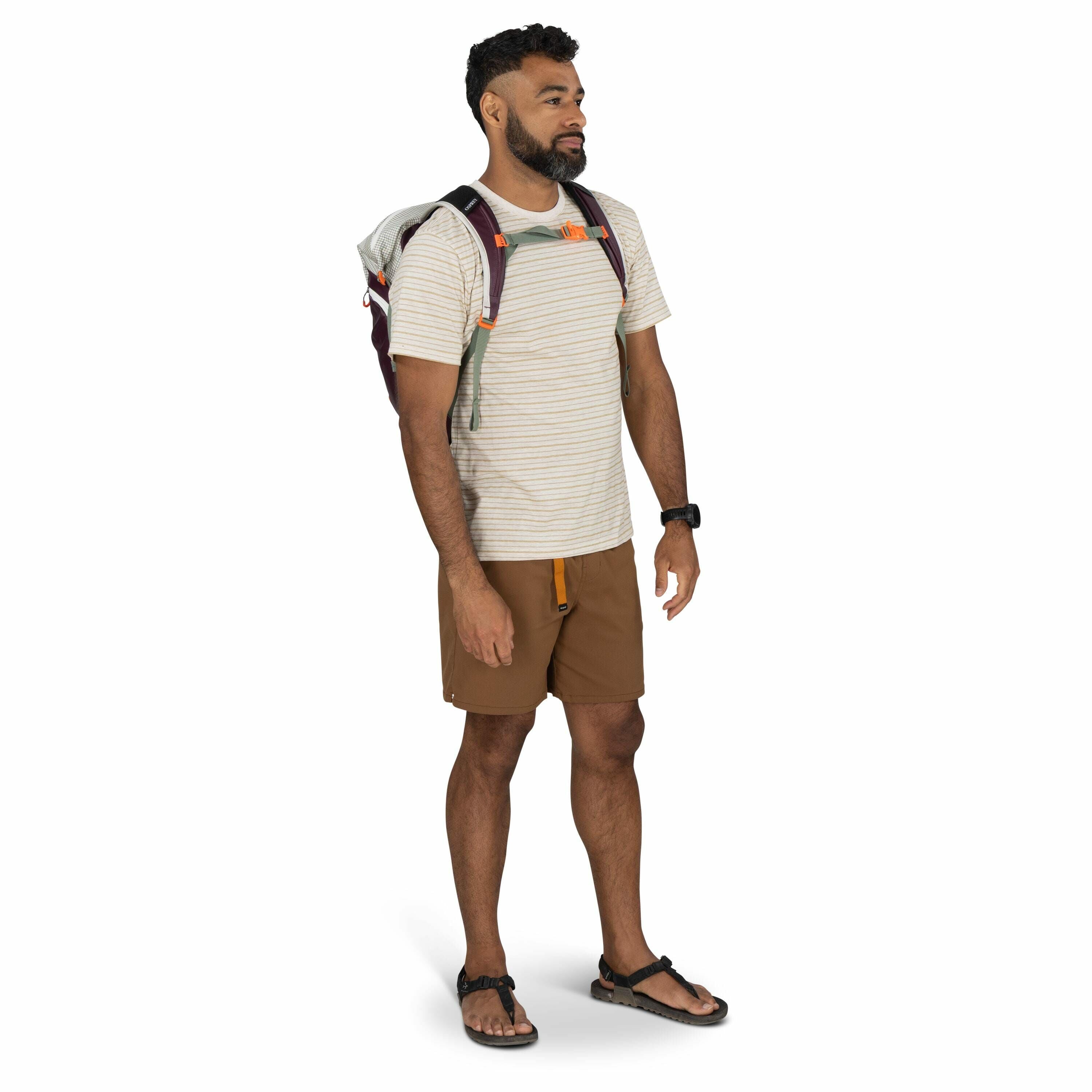 The image shows a man standing confidently, dressed in casual outdoor attire suitable for hiking or a day trip. He is wearing a light-colored, striped T-shirt paired with brown shorts, and he has a watch on his left wrist. On his feet, he sports black san