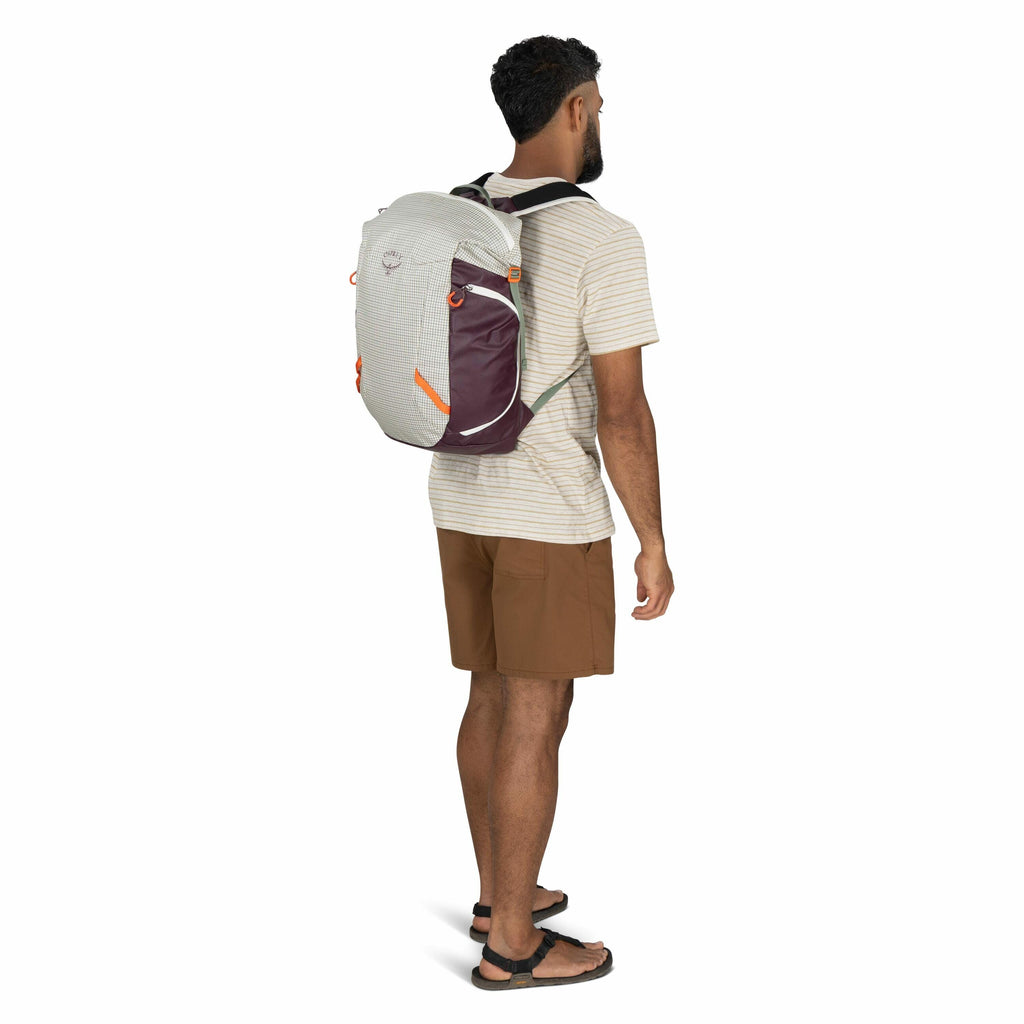 The image shows a man standing in a three-quarter profile view, facing slightly to the left. He is wearing a light-colored, short-sleeve striped T-shirt and brown shorts. On his back, he carries an Osprey backpack that is primarily light gray with maroon