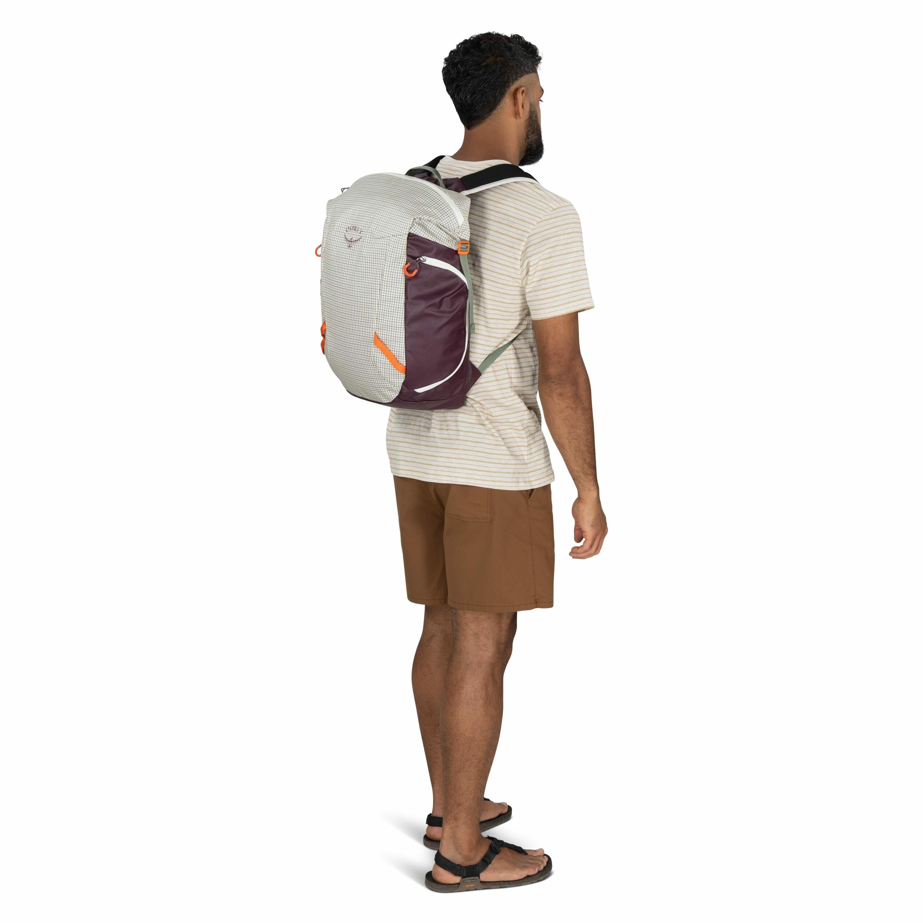 The image shows a man standing in a three-quarter profile view, facing slightly to the left. He is wearing a light-colored, short-sleeve striped T-shirt and brown shorts. On his back, he carries an Osprey backpack that is primarily light gray with maroon