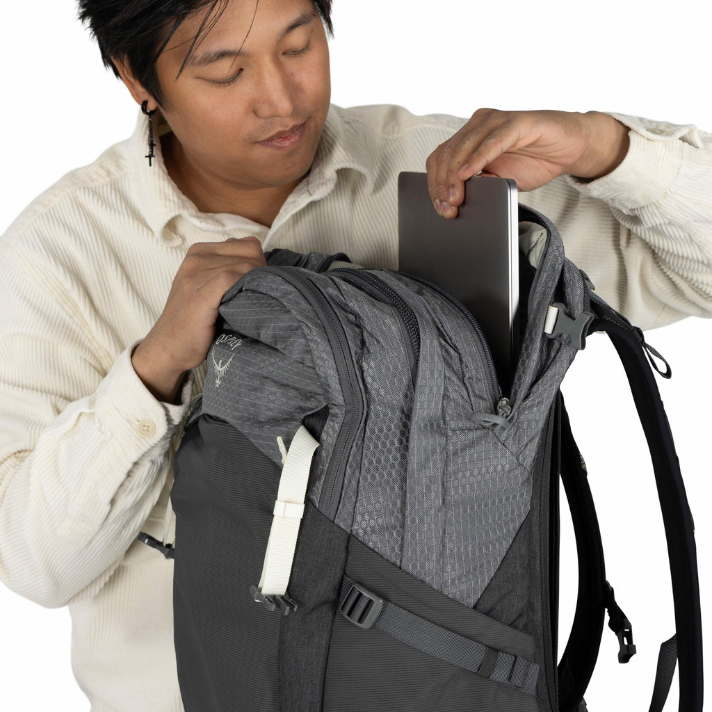 The image shows a person wearing a light-colored button-up shirt and inserting a laptop into a gray Osprey backpack. The backpack features a honeycomb pattern on the fabric and has a large zippered compartment where the laptop is being placed. There is a