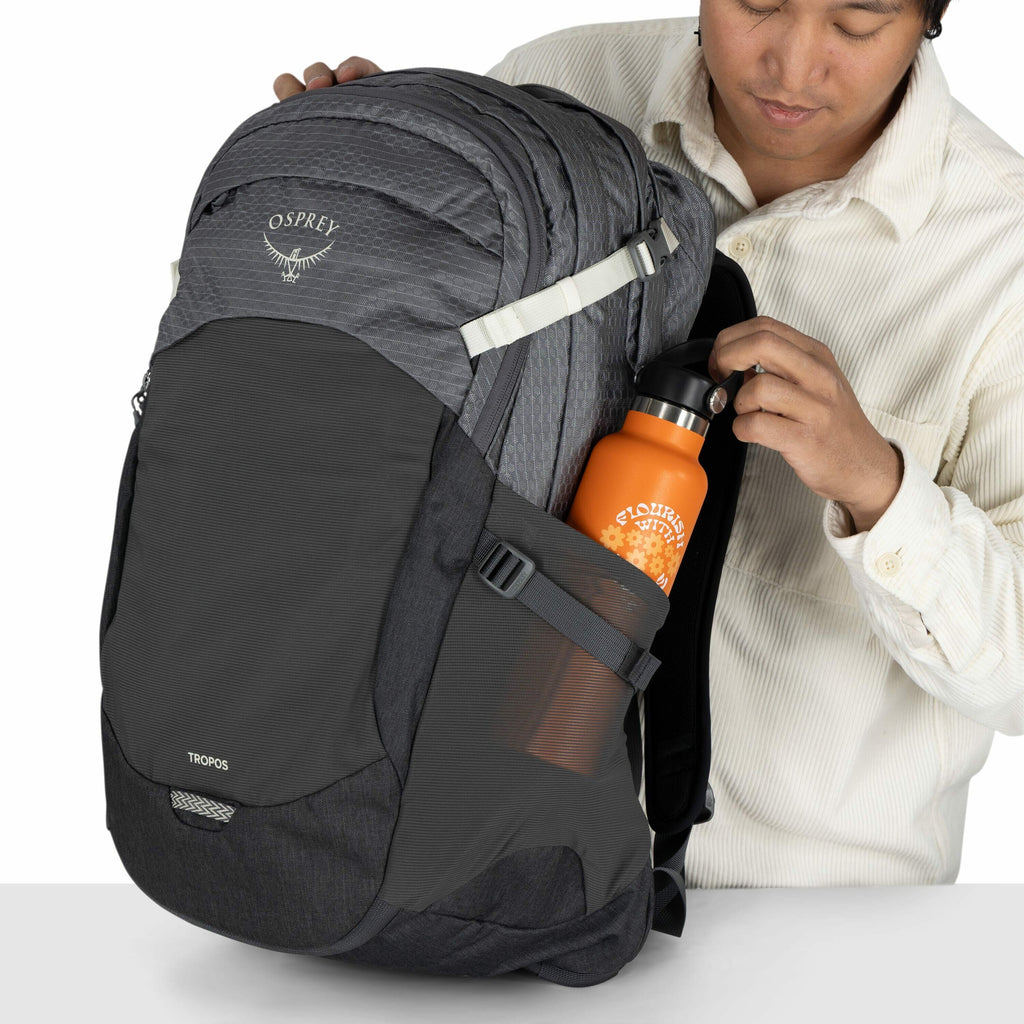 The image shows a person handling a large grey Osprey backpack, specifically the "TROPOS" model. The backpack has multiple compartments and is designed with white straps and a black mesh pocket on the side. The person is placing an orange water bottle int