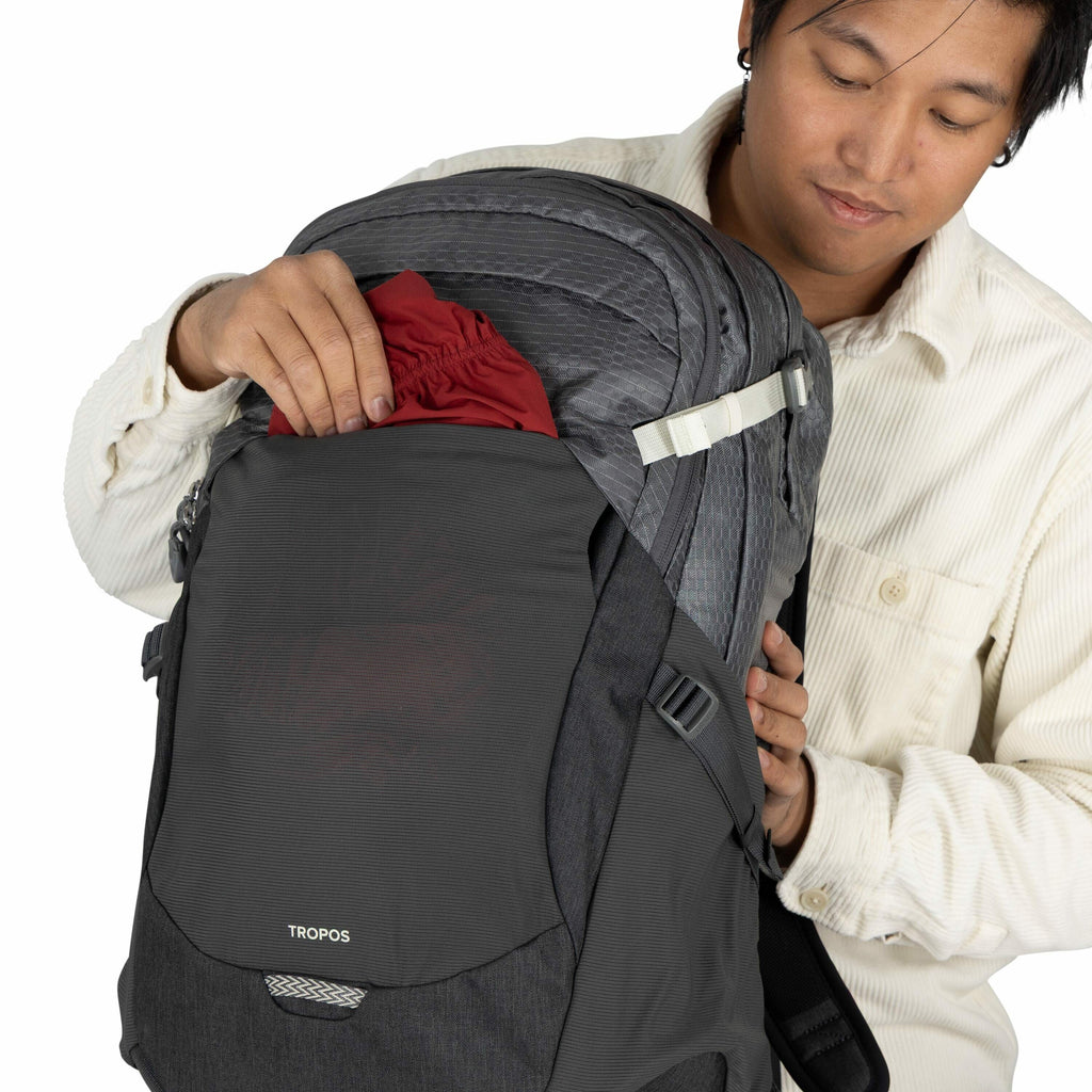 The image shows a person wearing a light-colored, long-sleeved button-up shirt, inserting a red fabric item into the front compartment of a dark gray backpack. The backpack features a design with various compartments, zippers, and adjustable straps. A log