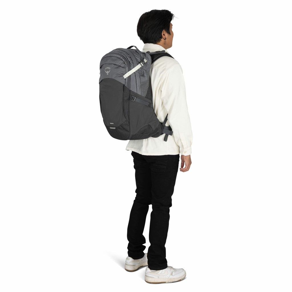 The image shows a person standing, turned slightly to the side, wearing a large, dark gray Osprey backpack. The backpack is designed with multiple compartments and a light gray pattern on its upper section. The person is dressed in a cream-colored shirt,