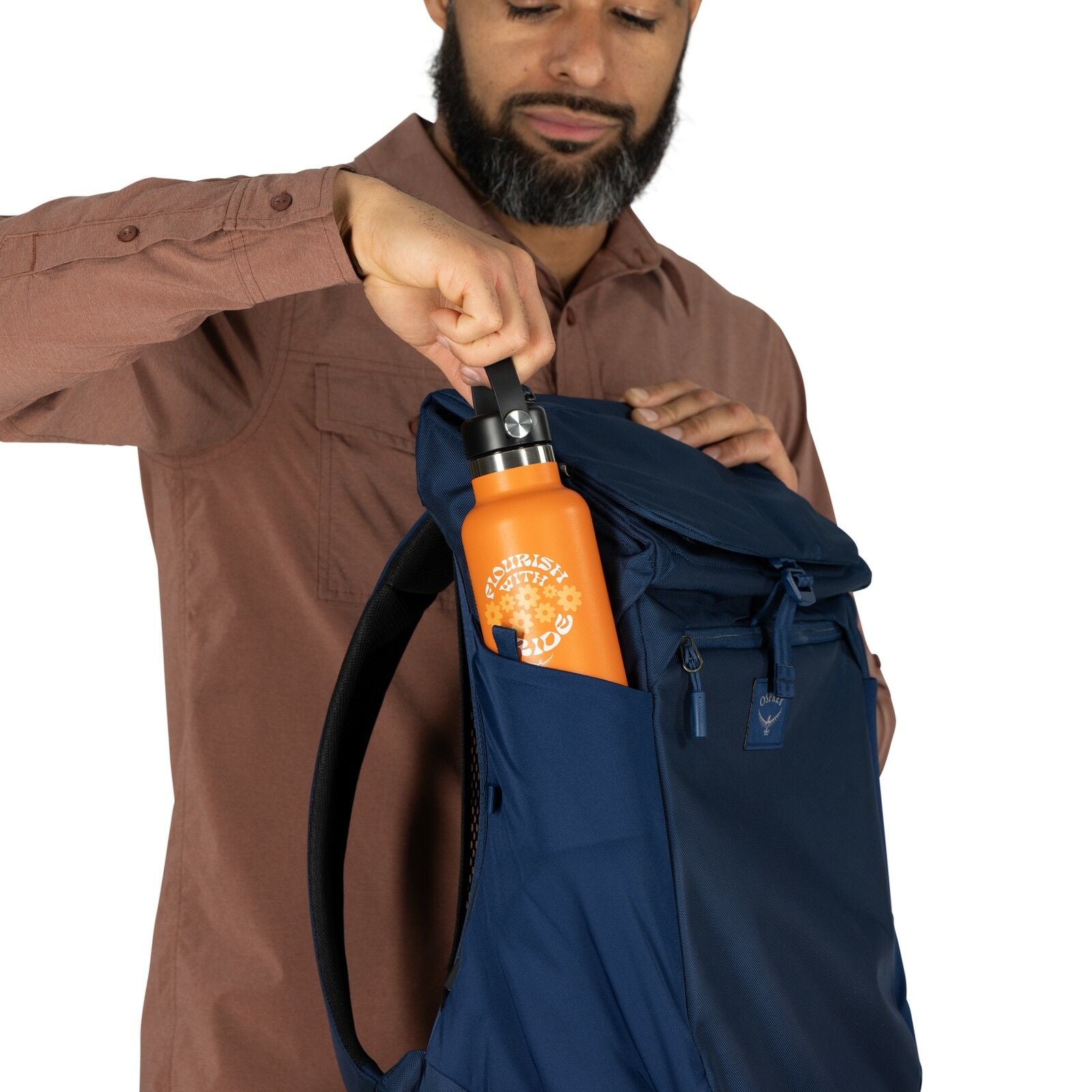 The image shows a person wearing a long-sleeved, button-up shirt in a shade of brown. They are placing or retrieving a bright orange water bottle from the side pocket of a dark blue backpack. The water bottle has a design on it featuring the phrase "CHARL