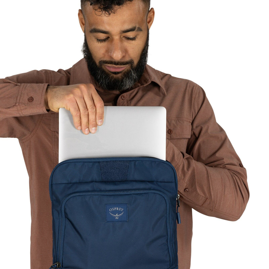 The image shows a person wearing a brown long-sleeved shirt, inserting or removing a silver laptop from a blue Osprey backpack. The backpack is held in front of the person, and the laptop is halfway into the main compartment of the backpack. The backpack