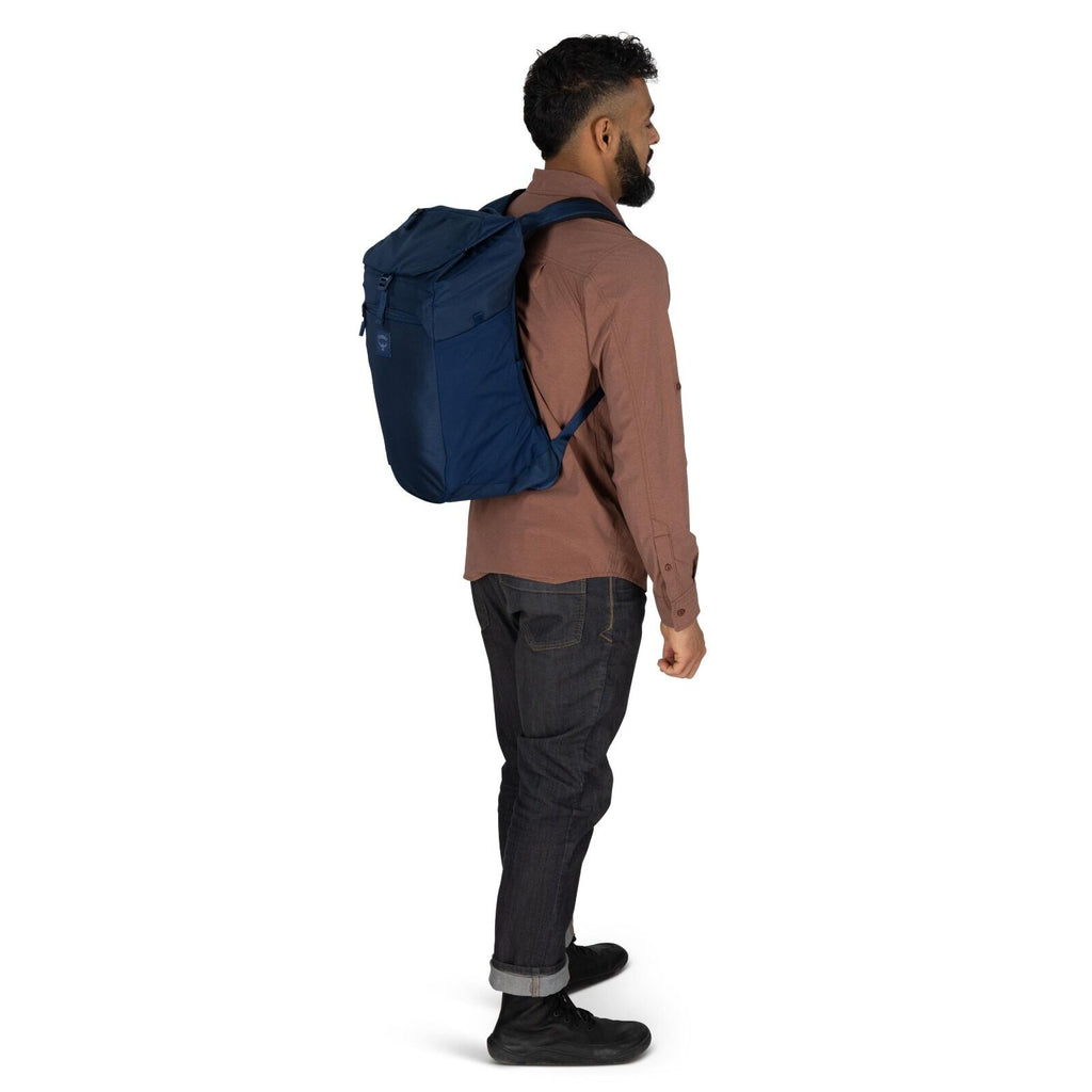 The image features a person standing in a profile view against a white and black background. The individual is wearing a brown long-sleeve button-up shirt, dark jeans, and black shoes. They are carrying a large blue backpack with several compartments, sug