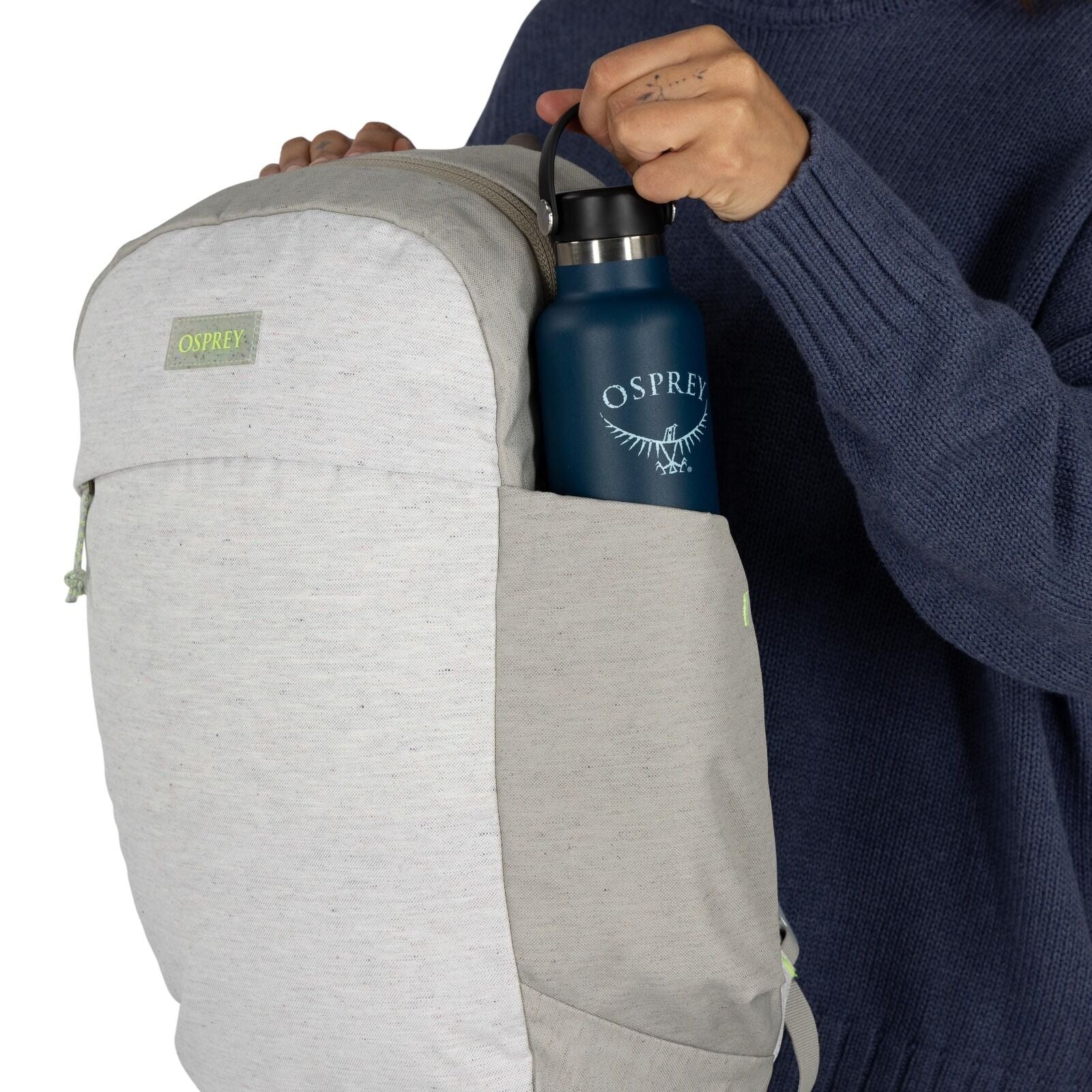 The image shows a person inserting a dark blue water bottle into a side pocket of a light gray backpack. The backpack and the water bottle are both branded with "Osprey," a company known for outdoor gear. The person is wearing a dark blue sweater, and the