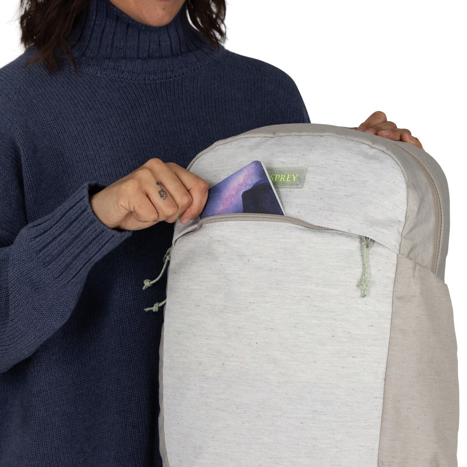 The image shows a person placing or retrieving a tablet or similar electronic device from the front pocket of a backpack. The backpack is primarily light gray, with a subtle, green "OSPREY" logo on the front. The person is wearing a dark blue, ribbed turt