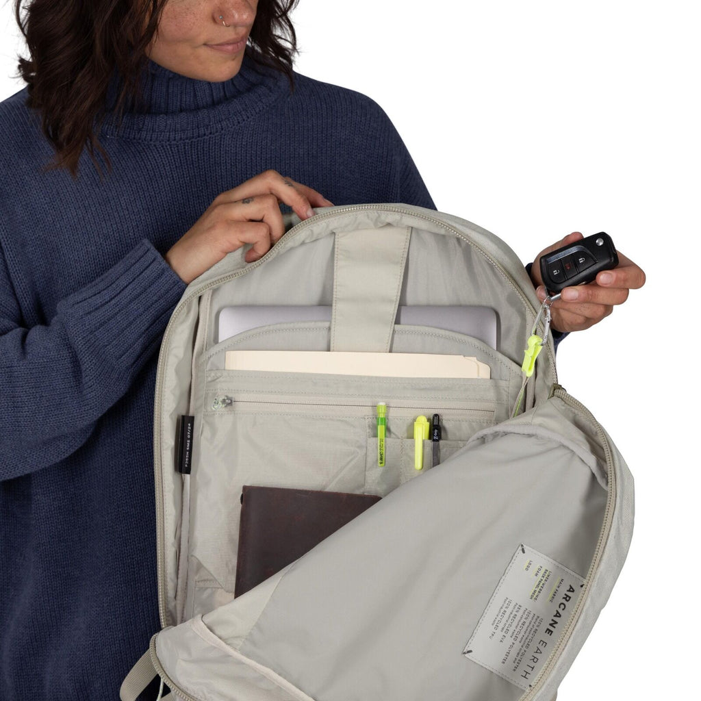 The image shows a woman holding open a light-colored backpack to reveal its contents. Inside the backpack, there is a laptop in a dedicated sleeve, folders, and several pens attached to an organizer panel. She is also holding a set of car keys, highlighti