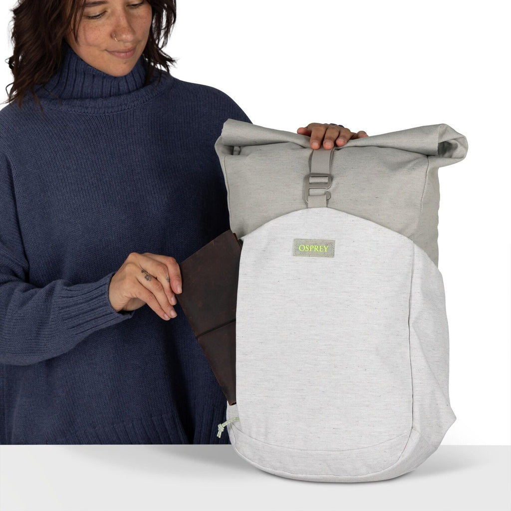 The image shows a person holding an Osprey backpack in front of them. The backpack is two-toned, with a light gray fabric on the bottom half and a darker gray on the top. It features a flap closure secured by a buckle, giving it a streamlined and practica