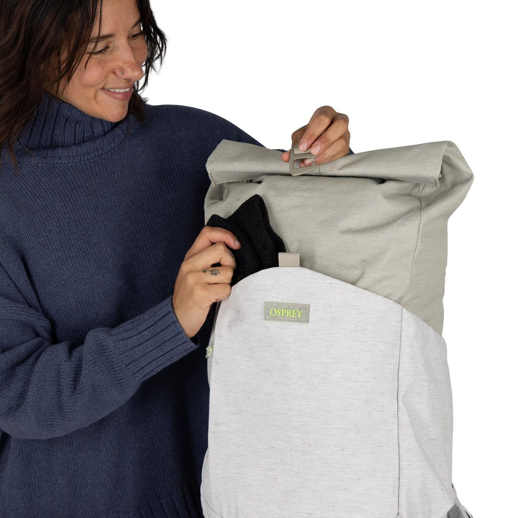 The image shows a person holding an Osprey backpack in front of them. The backpack is two-toned, with a light gray fabric on the bottom half and a darker gray on the top. It features a flap closure secured by a buckle, giving it a streamlined and practica
