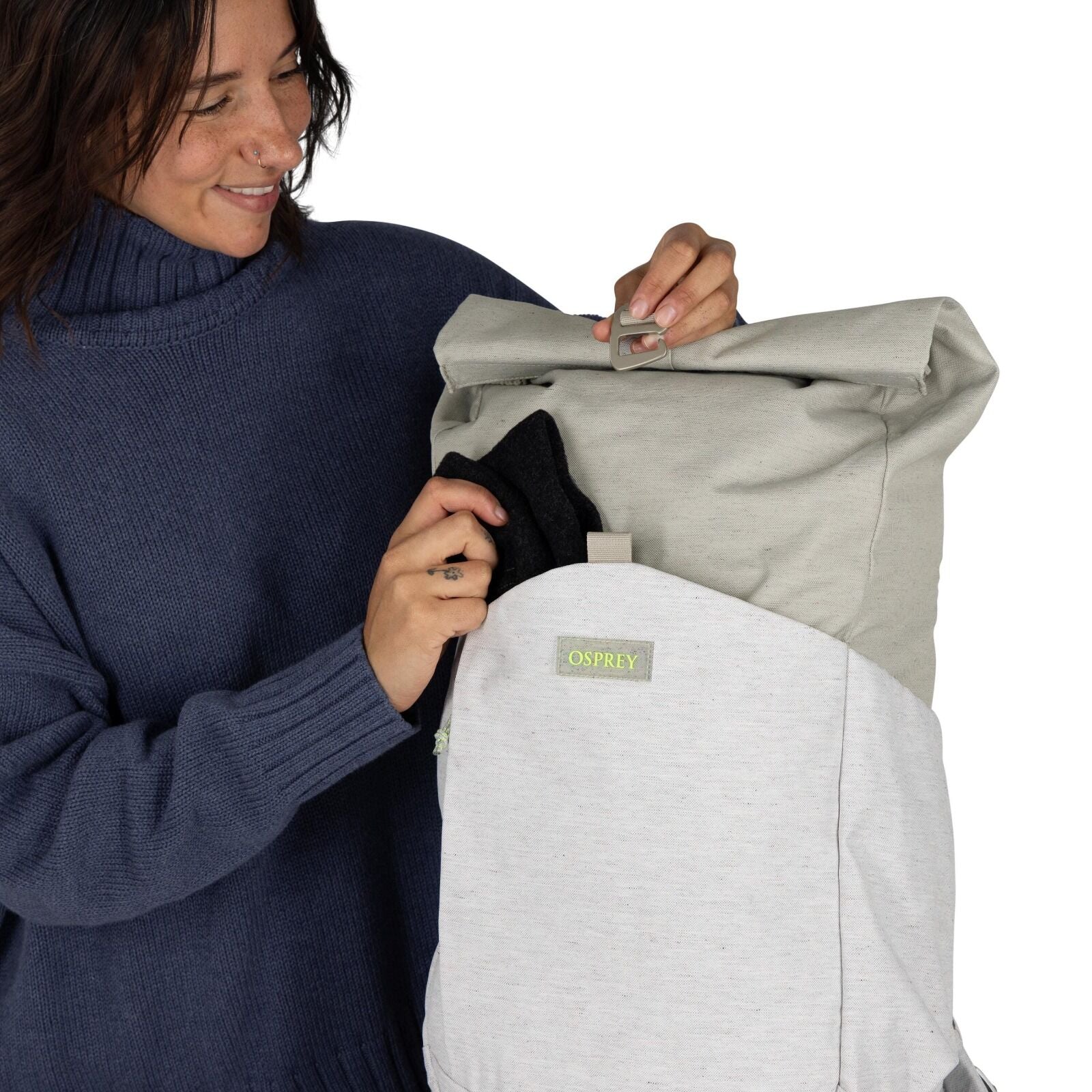 The image shows a person holding an Osprey backpack in front of them. The backpack is two-toned, with a light gray fabric on the bottom half and a darker gray on the top. It features a flap closure secured by a buckle, giving it a streamlined and practica